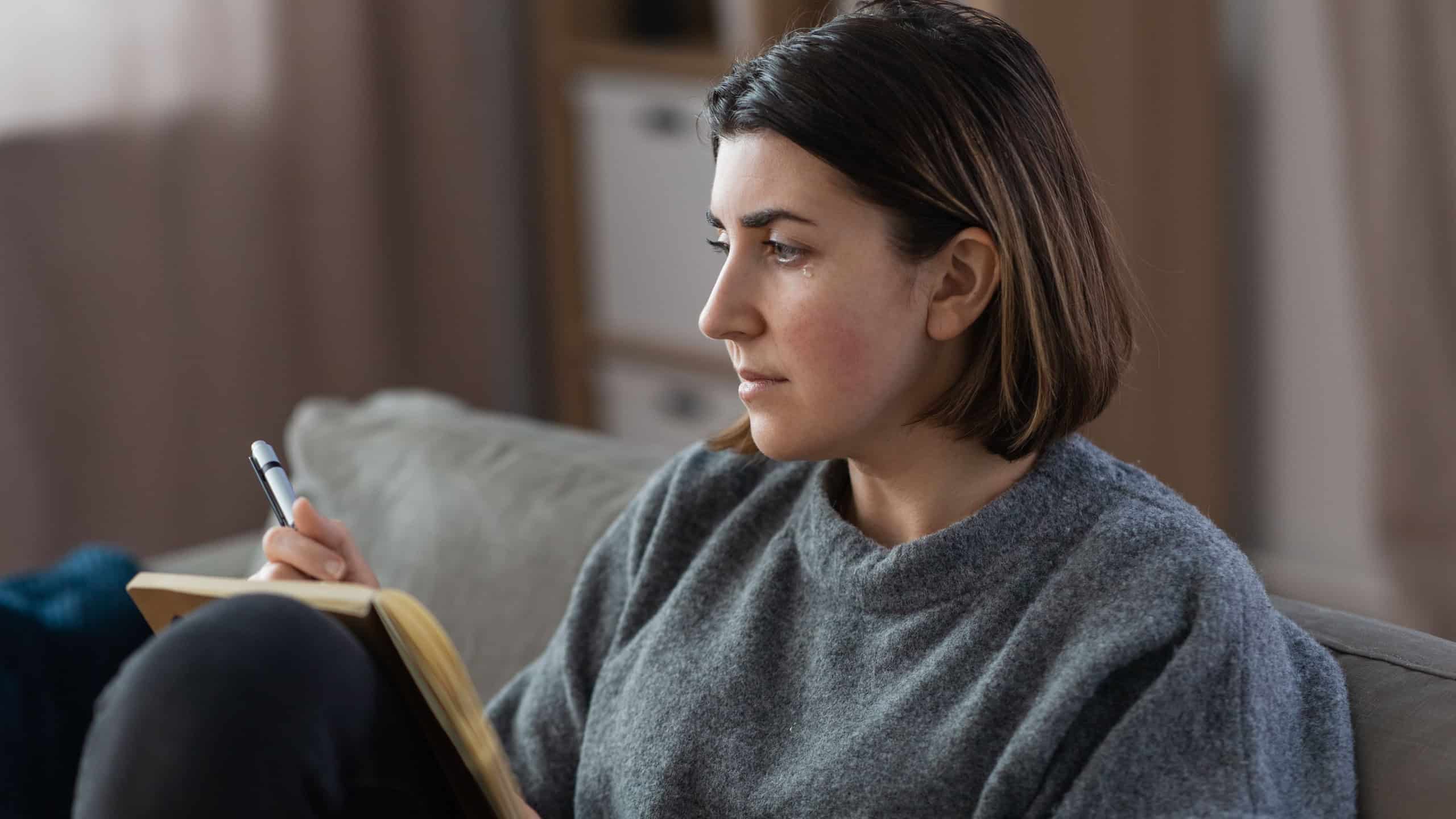 Stress, mental health and depression concept - sad crying woman with diary sitting on sofa at home