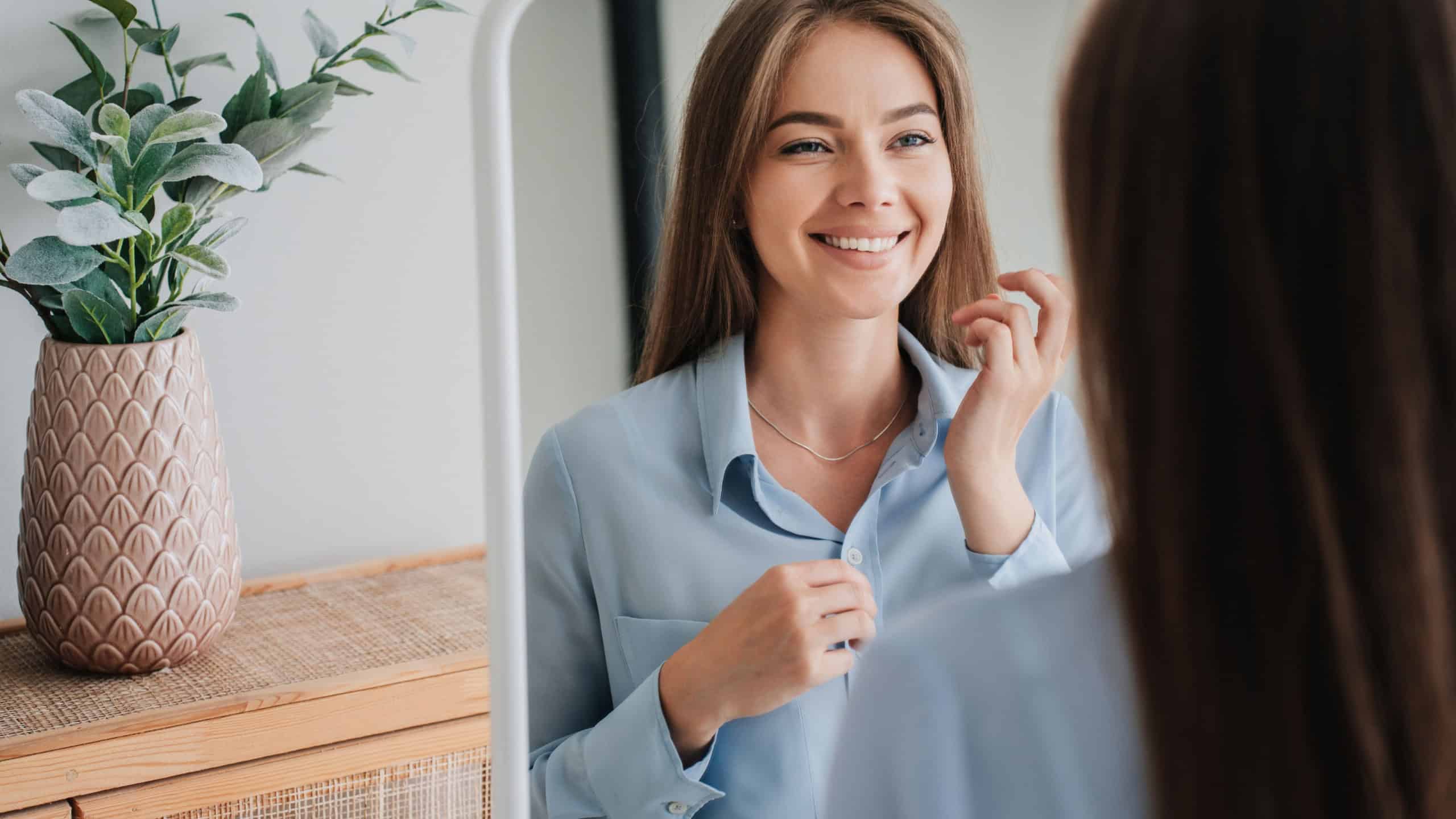 Cheerful young European woman in blue shirt standing at mirror toothy smiling satisfied by her skin health. Successful caucasian blonde woman toothy smiling at home, enjoying sunny morning at bedroom