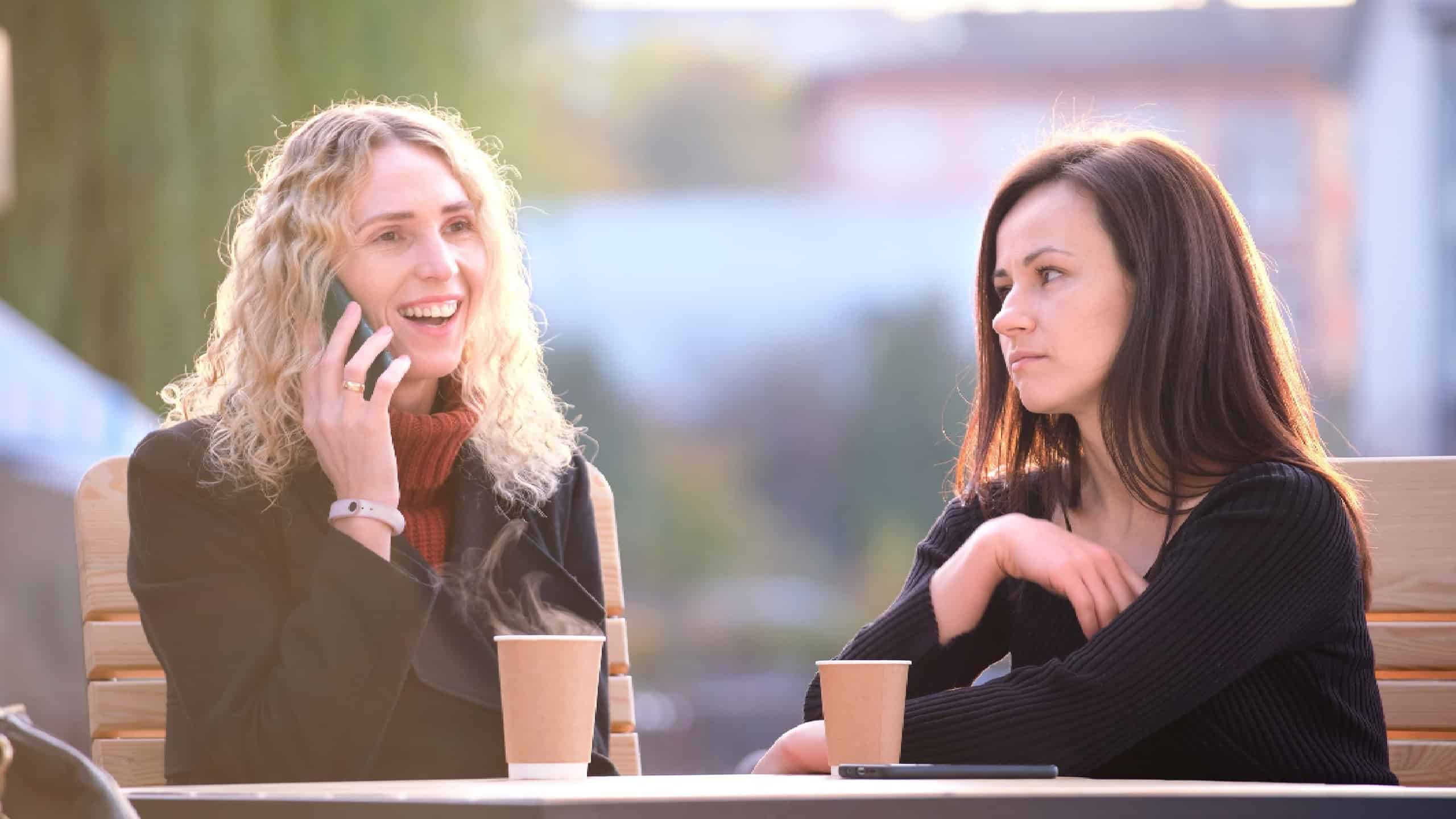 Happy young woman chatting on mobile phone ignoring her bored friend. Female friends sitting at street cafe outdoors and having hard time communicating with each other. Friendship problems concept