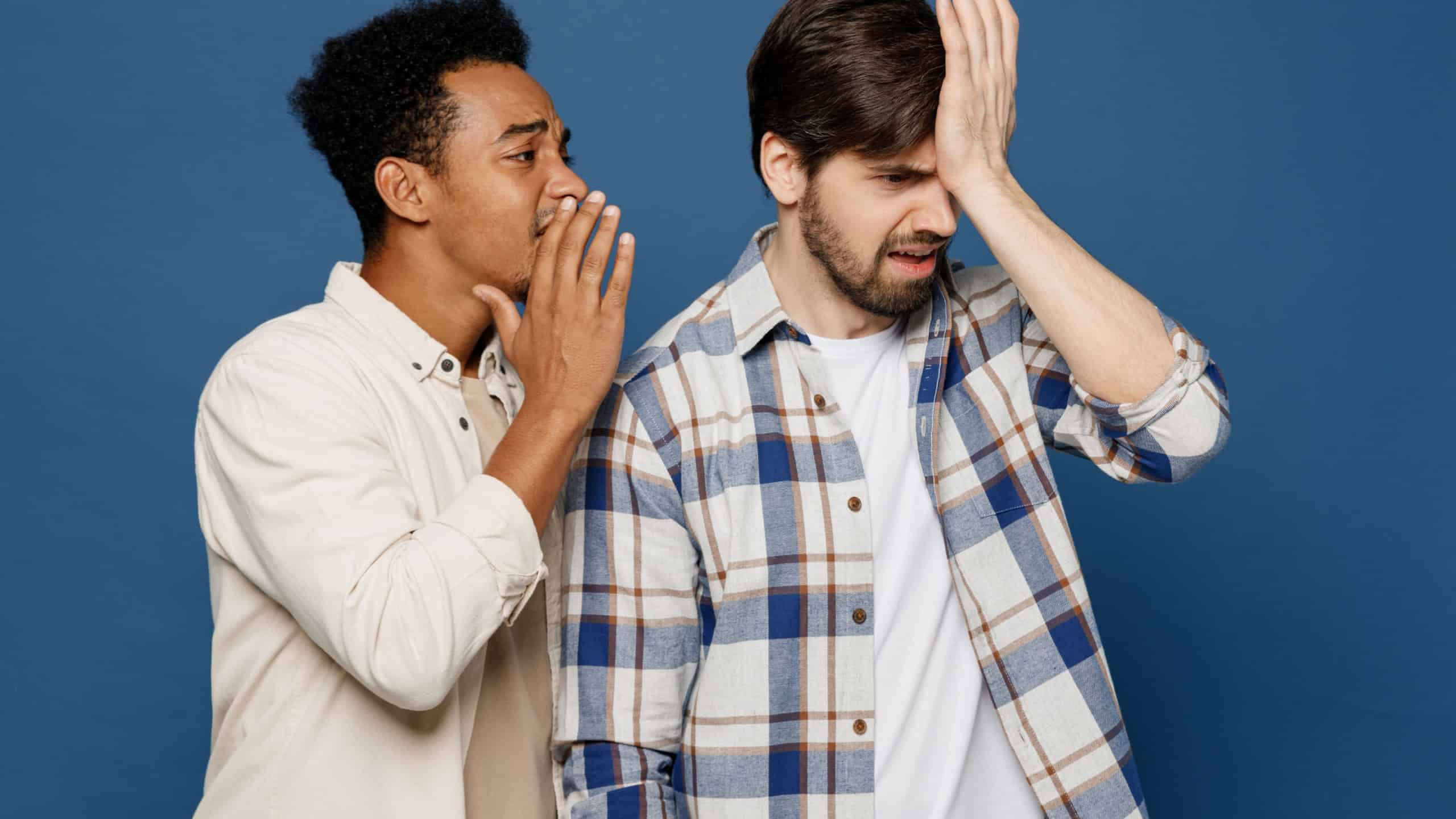 Young two friends shocked sad men wear white shirts together whisper gossip and tells secret behind his hand sharing news show isolated plain dark royal navy blue background. People lifestyle concept