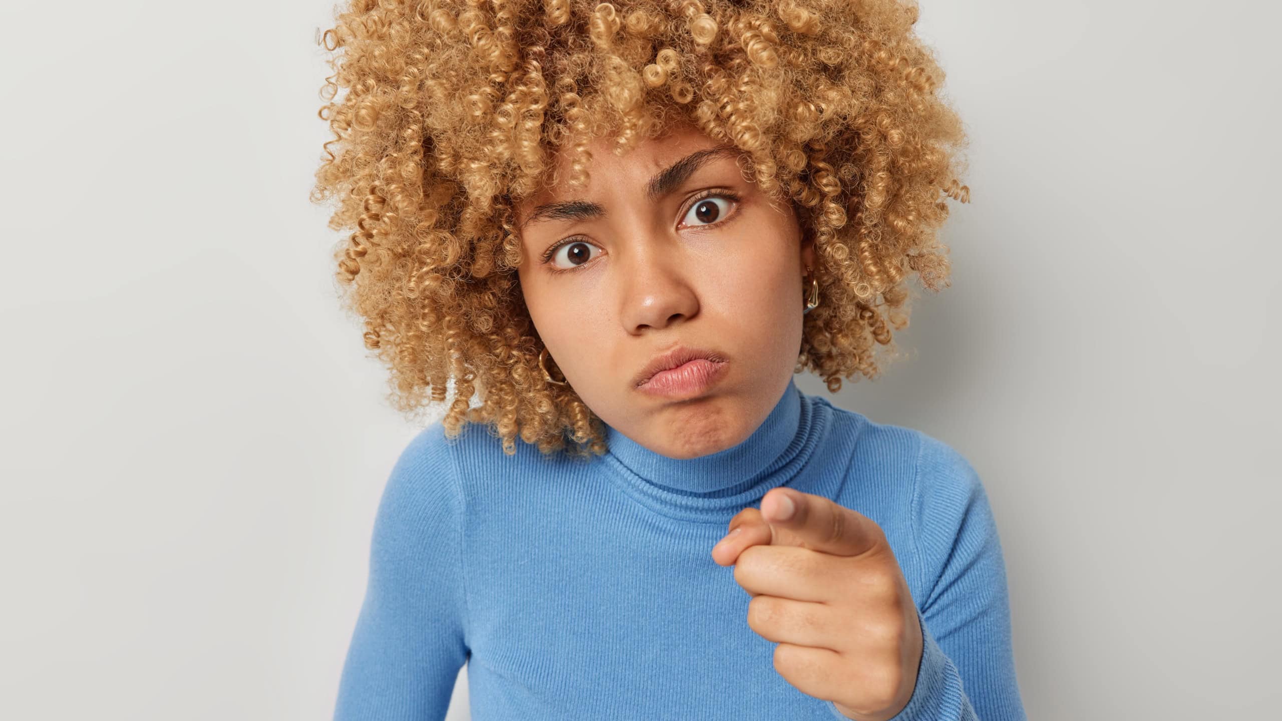 Portrait of serious curly haired woman points index finger at camera blames you and feels angry dressed in blue turtleneck isolated over grey background. Displeased female model indicates at camera