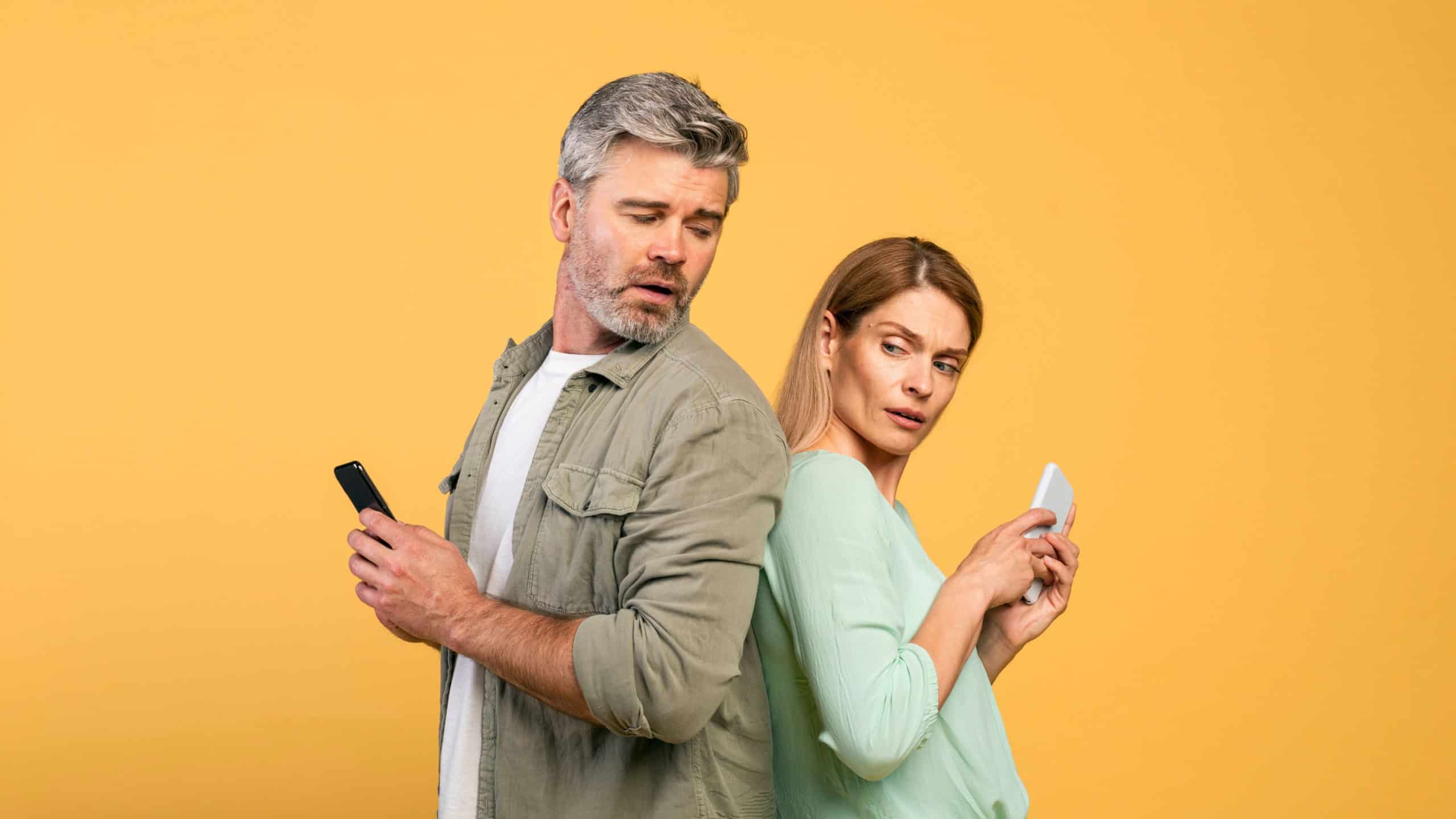Privacy concept. Middle aged caucasian man trying to look at his wife's smartphone, woman hiding screen of her gadget, standing over yellow studio background