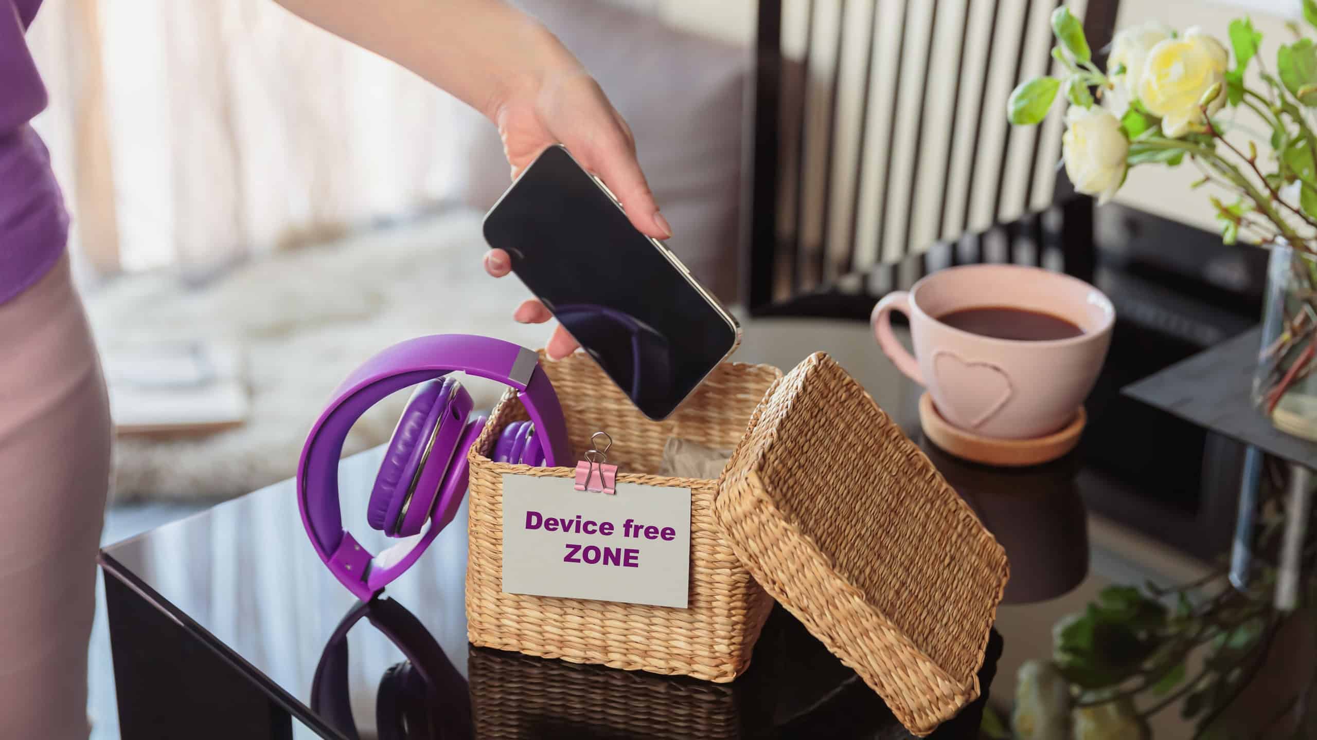 Close up of hand holding smartphone in wicker basket with inscription Device free zone. Woman putting phone into box with different gadgets at home. Digital detox and technology dependance concept