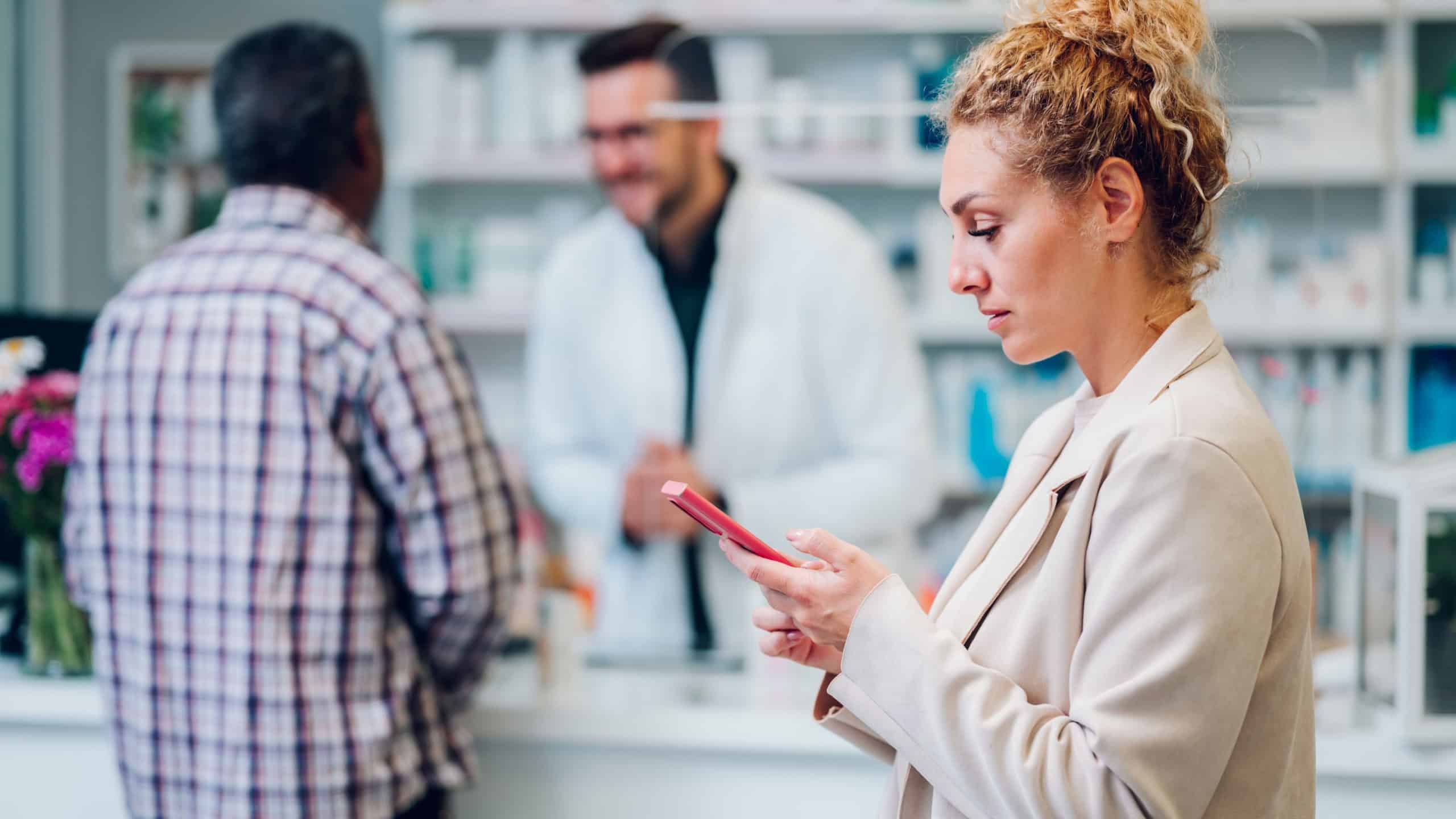 Portrait of a middle age woman patient customer shopping at pharmacy and using a smartphone. A queue of people in a drugstore, standing behind each other waiting to be served by the pharmacist.