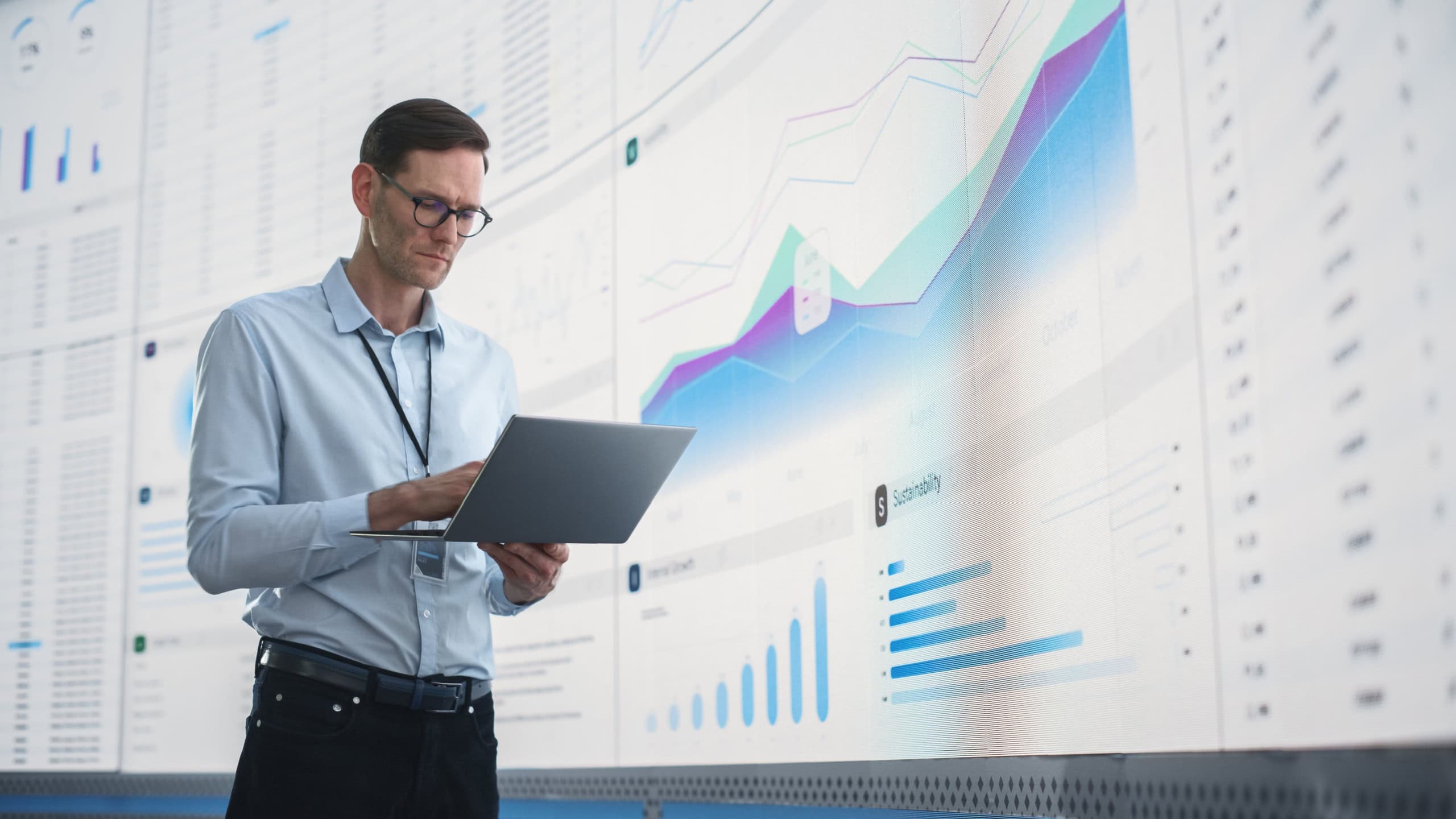 In The Office: Male Monitoring Specialist Holding Laptop Computer And Standing Next To Big Digital Screen With Graphs And Charts. Caucasian Man Analyzing Data, Performance Records, And Filing Report.