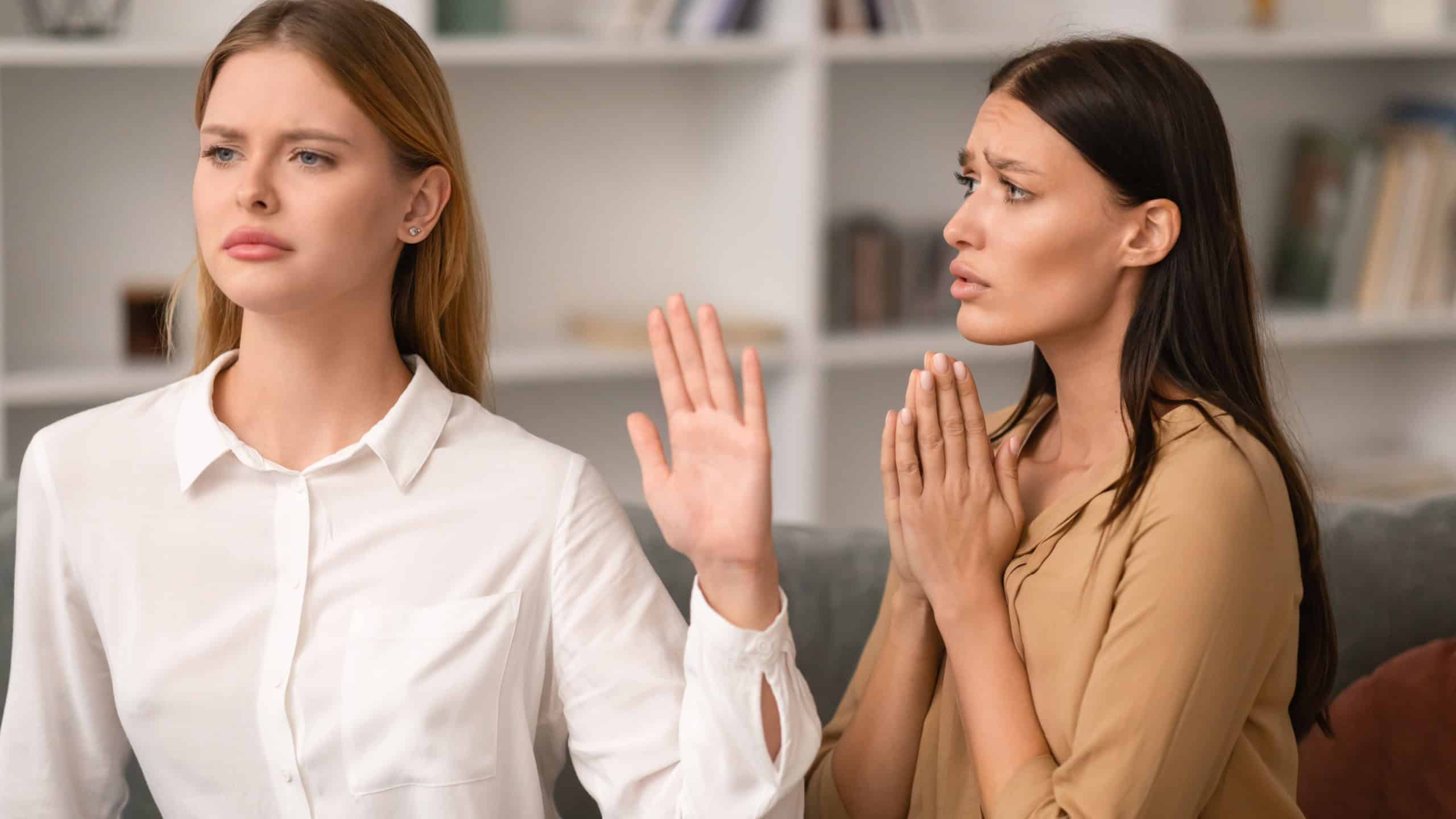 Woman Saying No and Gesturing Stop In Refusal To Friend Begging For Favor Or Forgiveness After Quarrel, Sitting On Couch In Modern Living Room Interior. Friendship Relationship Problems