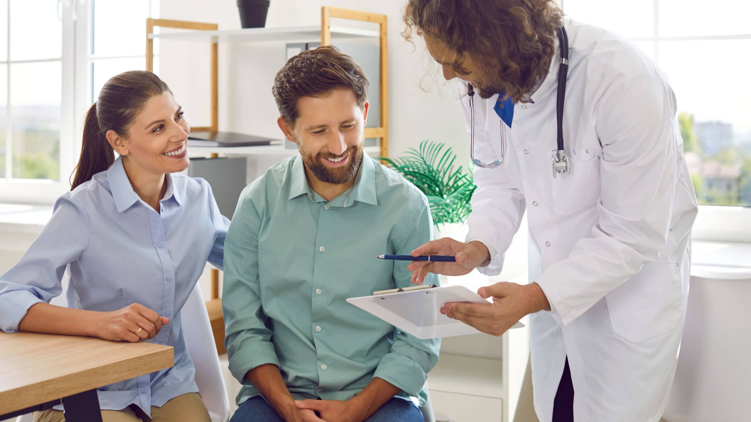 Friendly gynecologist advises couple on pregnancy and shows them clipboard with test results. Smiling positive Caucasian couple planning pregnancy consults in modern clinic of reproductive medicine.