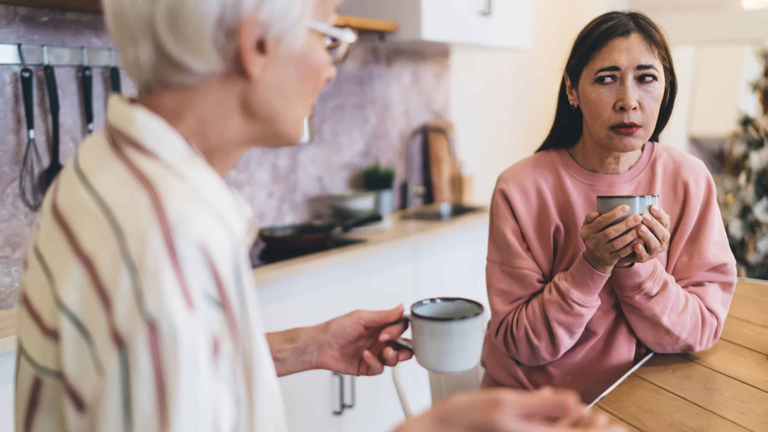 Skeptical senior female having conversation with girlfriend while drinking hot beverage discussing news in modern kitchen spending time together at home