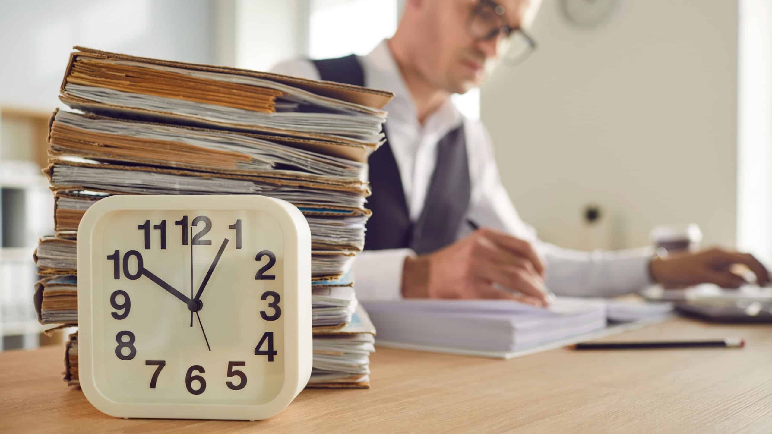 Stack of papers, table clock and businessman engrossed in work, symbolize the overtime and value of time. Image showing dedication, hard work, business environment and importance of time management.