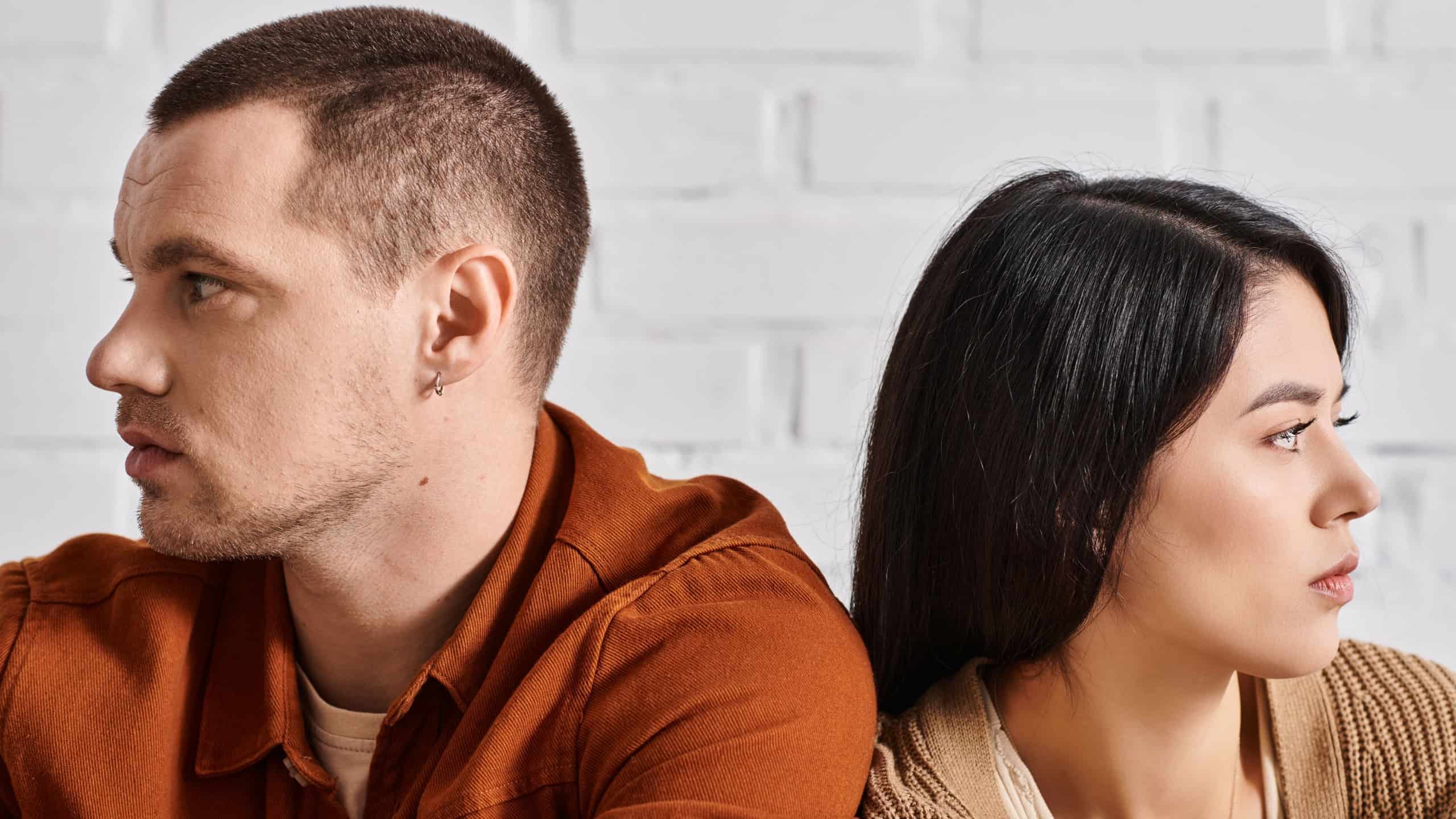 offended interracial couple sitting back to back and looking away in living room, divorce concept