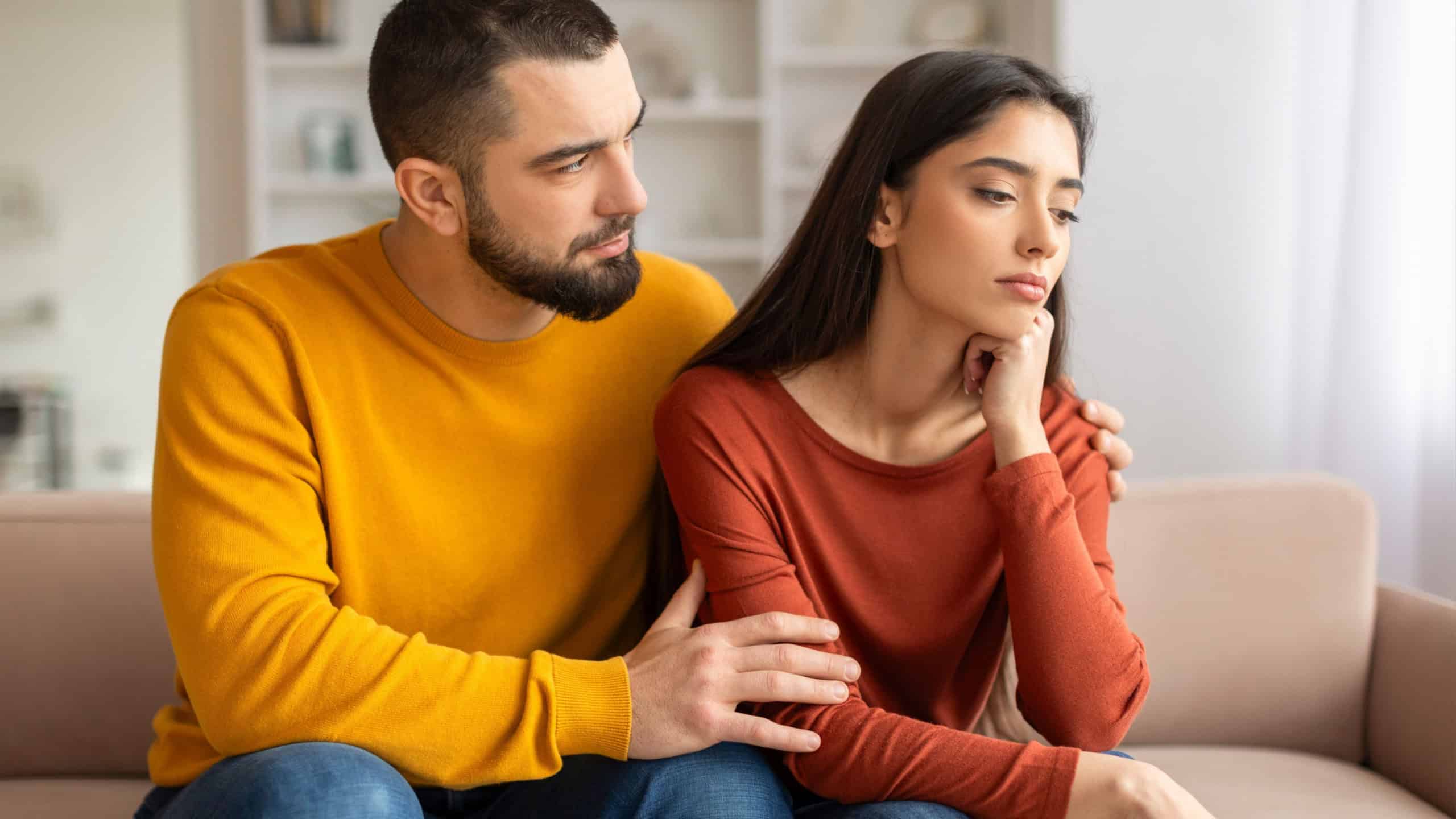 Compassionate young man comforting his upset wife at home, loving husband providing support and expressing empathy while sitting together on couch in living room interior, closeup shot