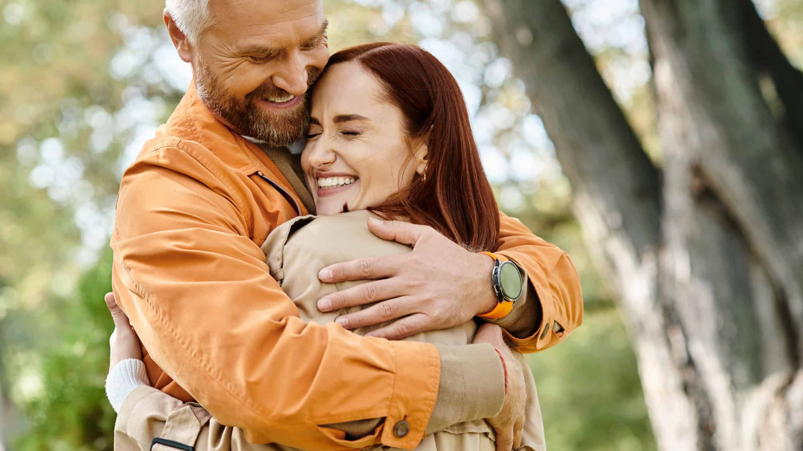 A man tenderly hugs a woman in a park filled with lush greenery.