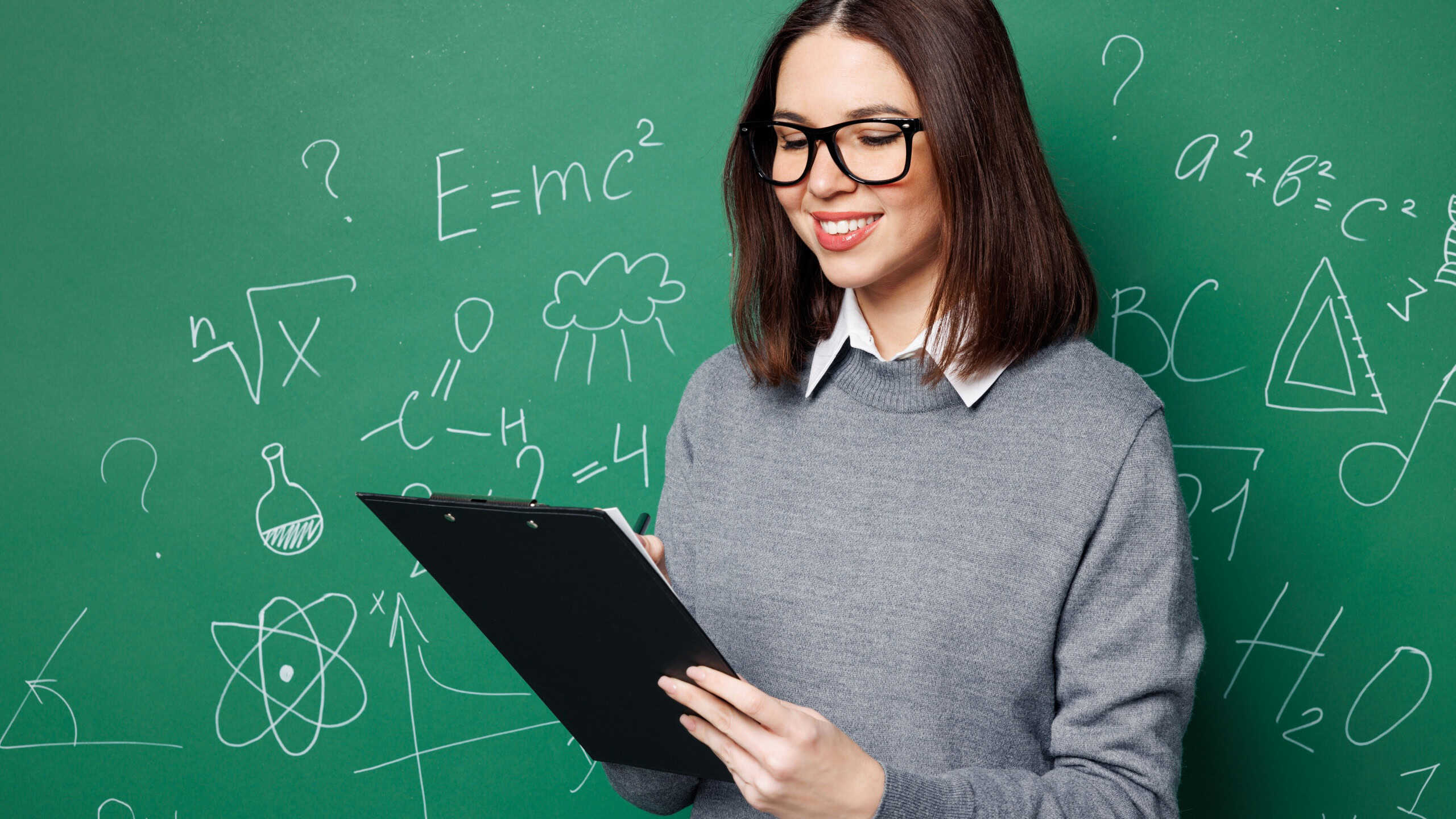 Young smart woman wearing grey casual shirt glasses clipboard with paper documents write down notes isolated on green wall chalk blackboard background. Education in high school college concept