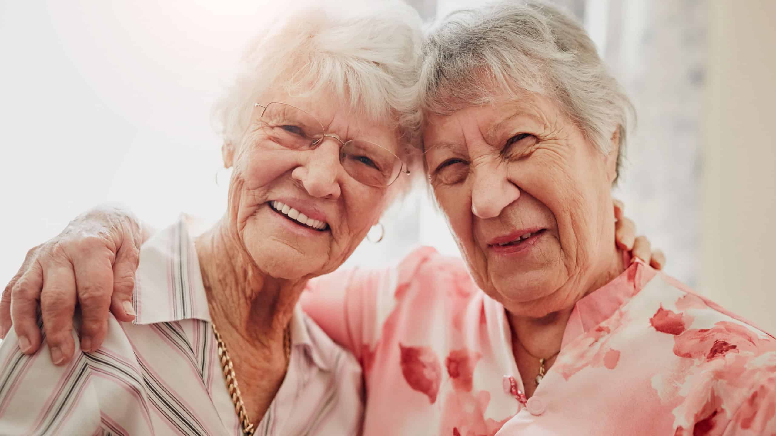 Elderly women, friends and hug in house with smile in portrait for care, love or bonding with reunion. Senior people, embrace and happy for connection in morning, lounge or together in nursing home