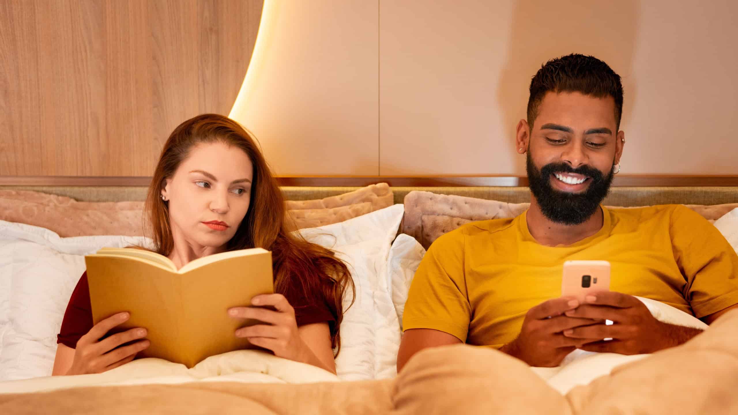 Unhappy woman in bed while her partner uses a cell phone during the evening. Distracted man enjoying his smartphone while his partner looks displeased in a cozy bedroom settin