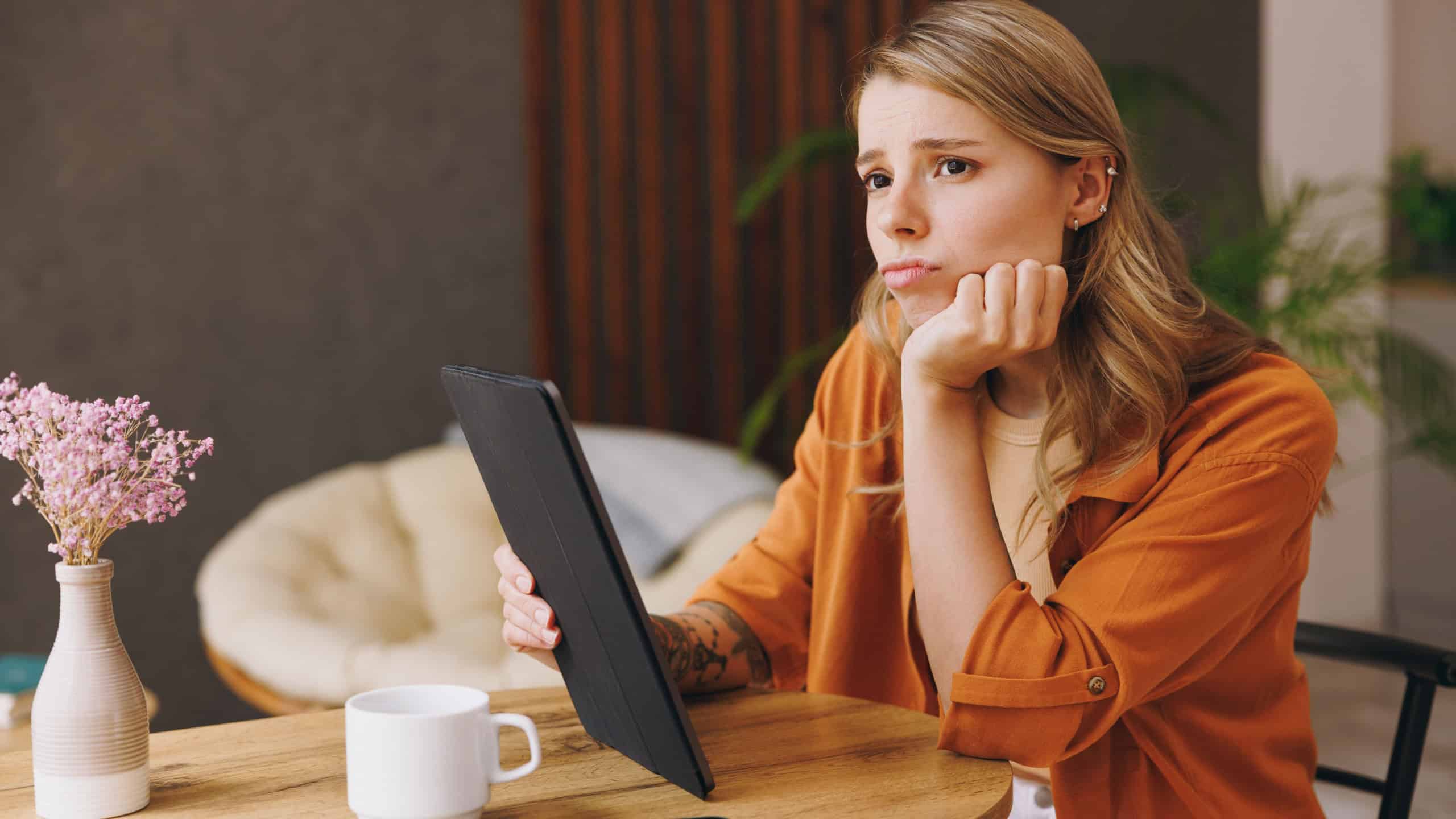 Young sad IT woman wear orange shirt hold use digital tablet pc computer sit alone at table in coffee shop cafe relax rest at restaurant in free time indoors. Freelance mobile office business concept