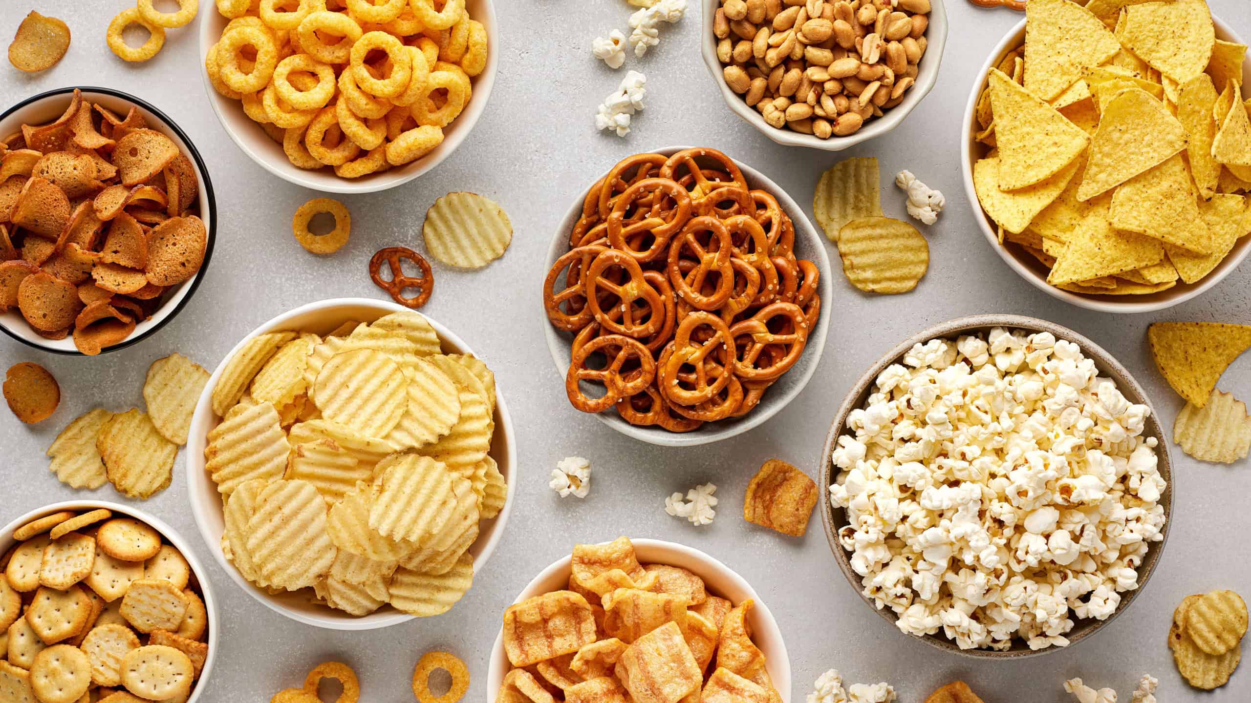 Set of many bowl with different salty snacks on light gray background. Chips, popcorn, nuts, nachos and pretzels. Party food.