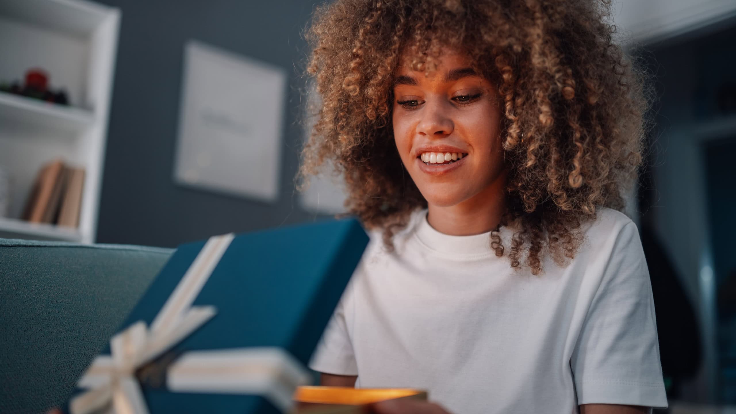 Excited woman with curly hair opens gift box, smiling in anticipation. Blue and gold ribbon hints at special occasion. Captures joy of giving and receiving in cozy setting