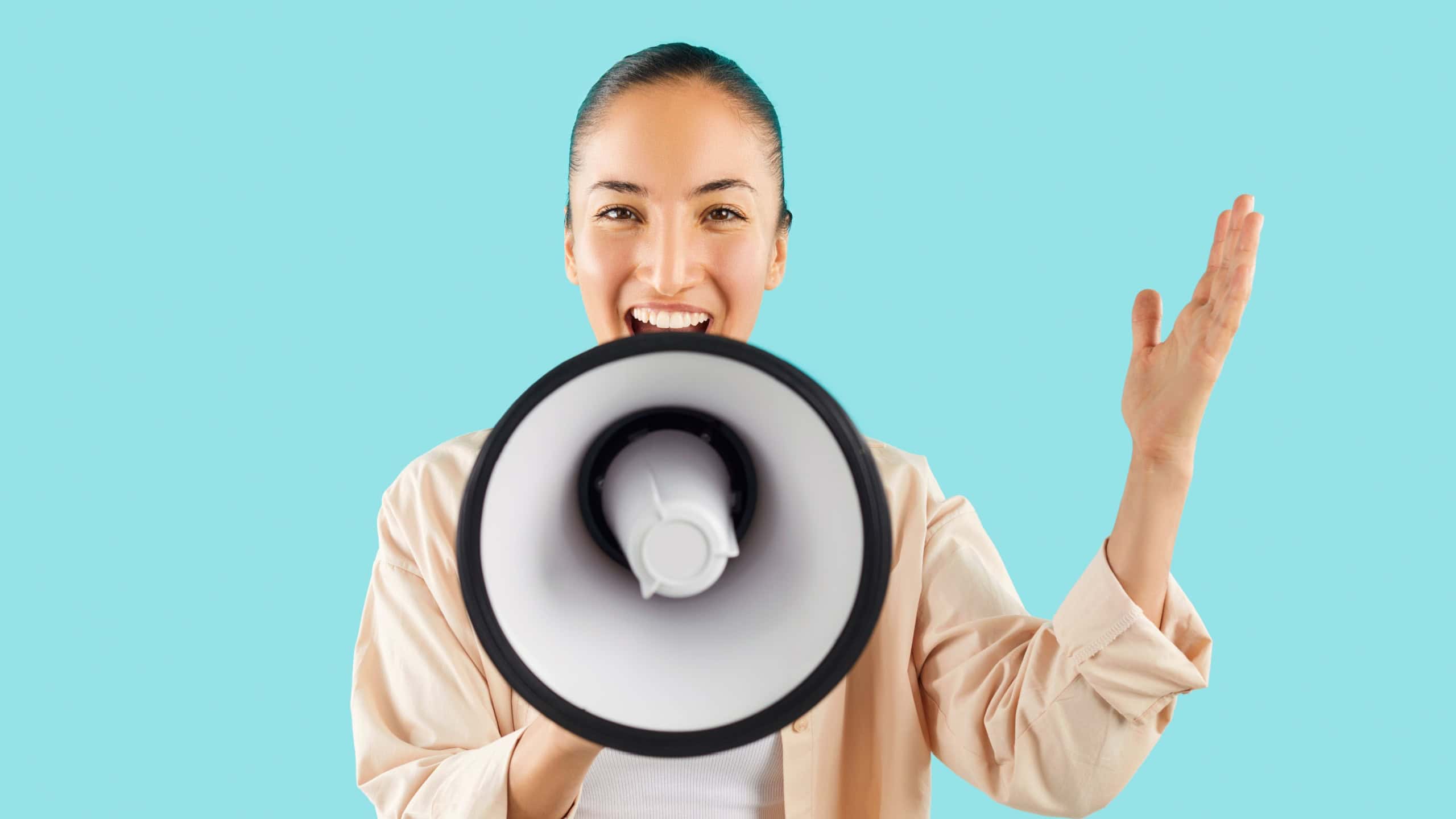 Studio shot of happy young Asian woman screaming in megaphone. Cheerful young girl isolated on bright blue background holding loud speaker in hand, looking at camera and talking with amplified voice
