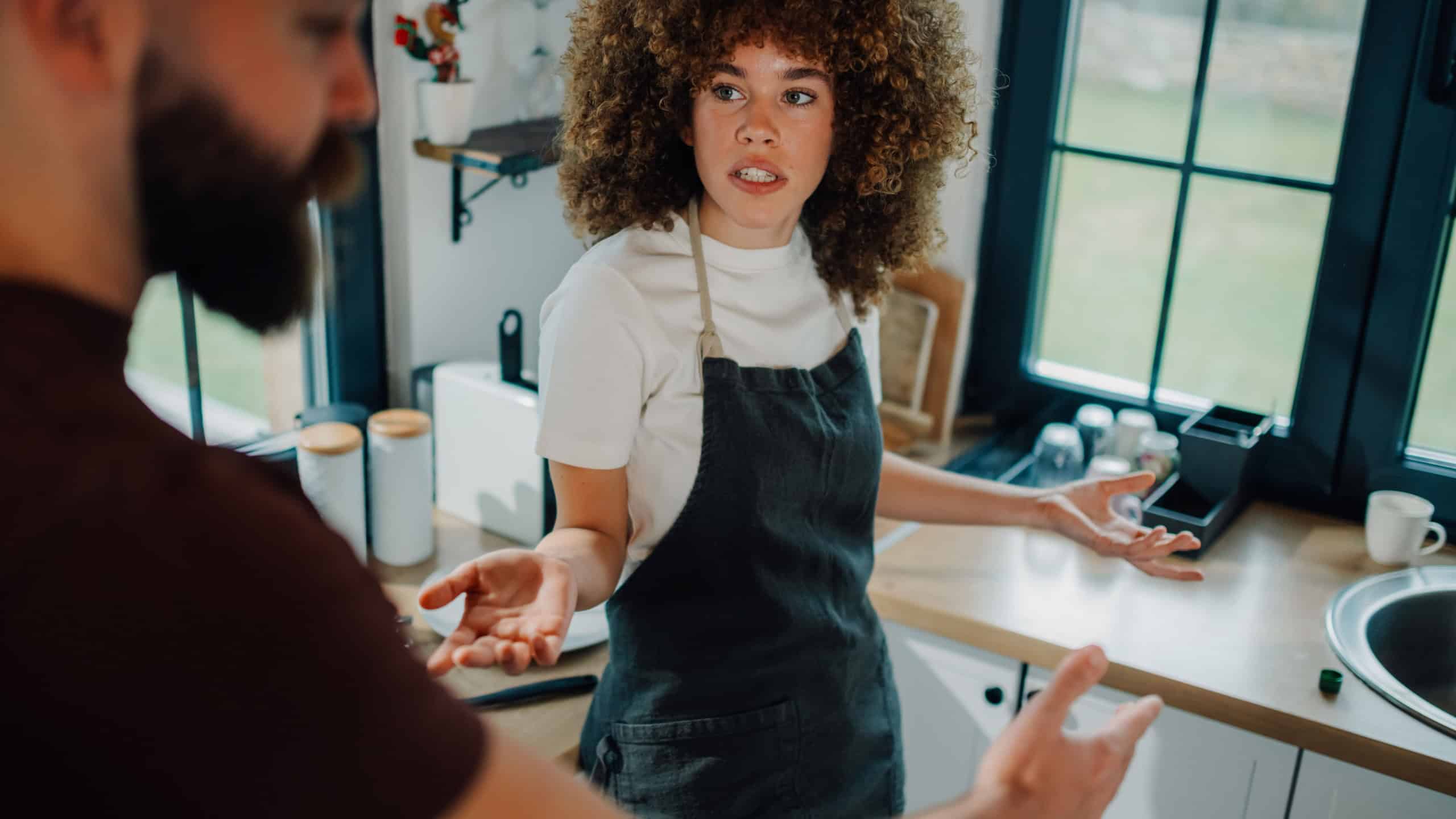 Young frustrated woman wearing apron arguing with her husband at home in kitchen, complaining about household chores and responsibilities