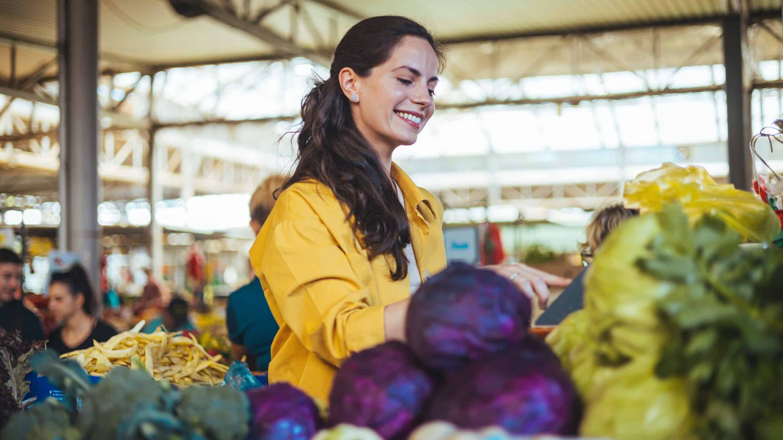 A cheerful woman in a vibrant yellow shirt at a market stall displaying fresh vegetables, fostering a sense of local community and healthy living.