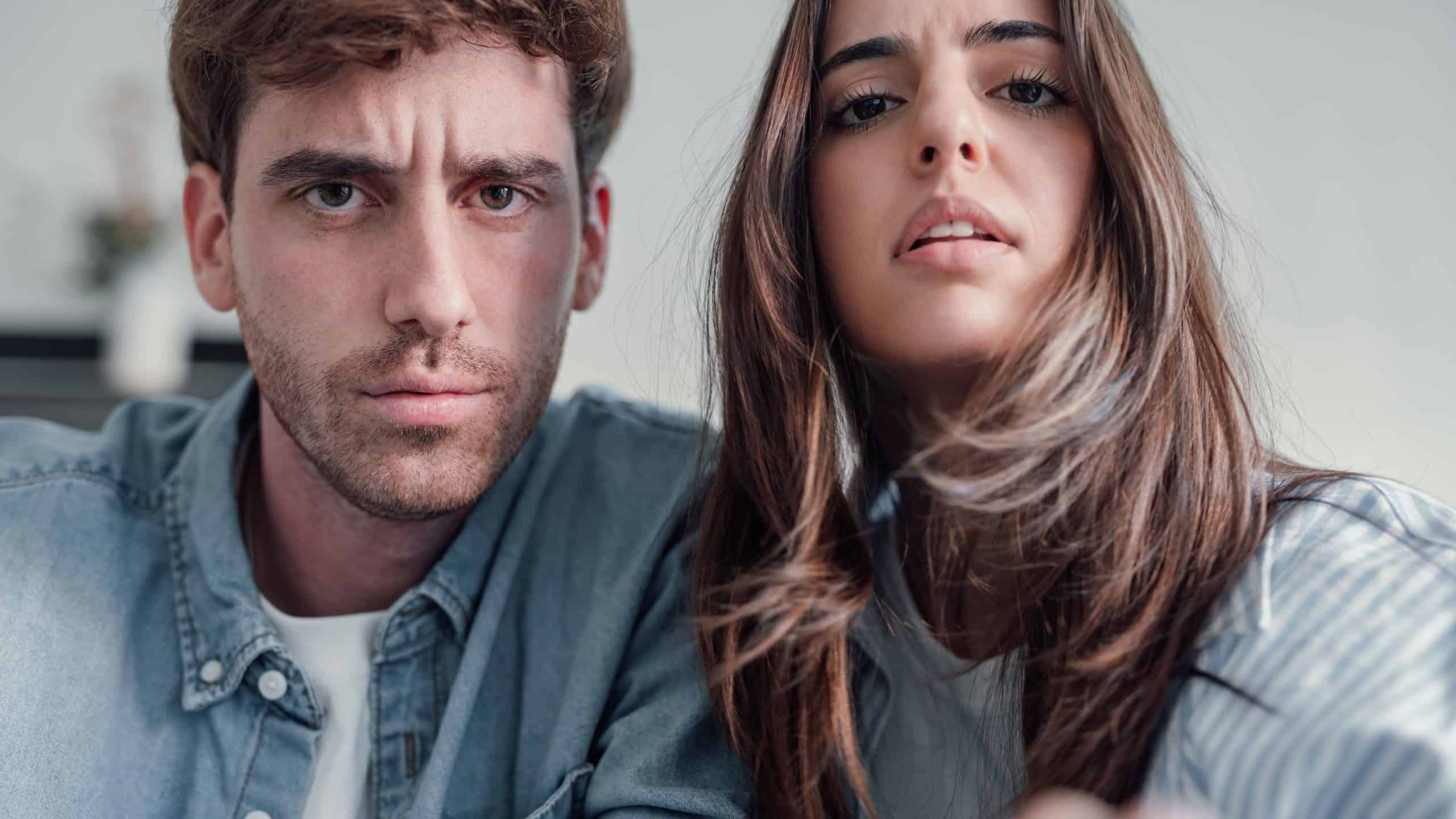 Confused couple touching camera clicking screen thinking worried doubting if click. Puzzled perplexed young two people with distrustful face unbelieving suspicious looking at home on sofa