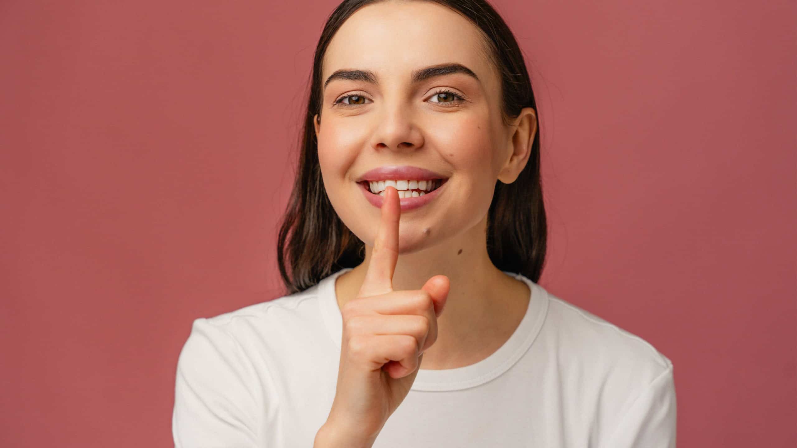 A cheerful individual holds a finger to their lips in a soft silence gesture against a pastel pink backdrop. The scene portrays happiness, peace, and playfulness.