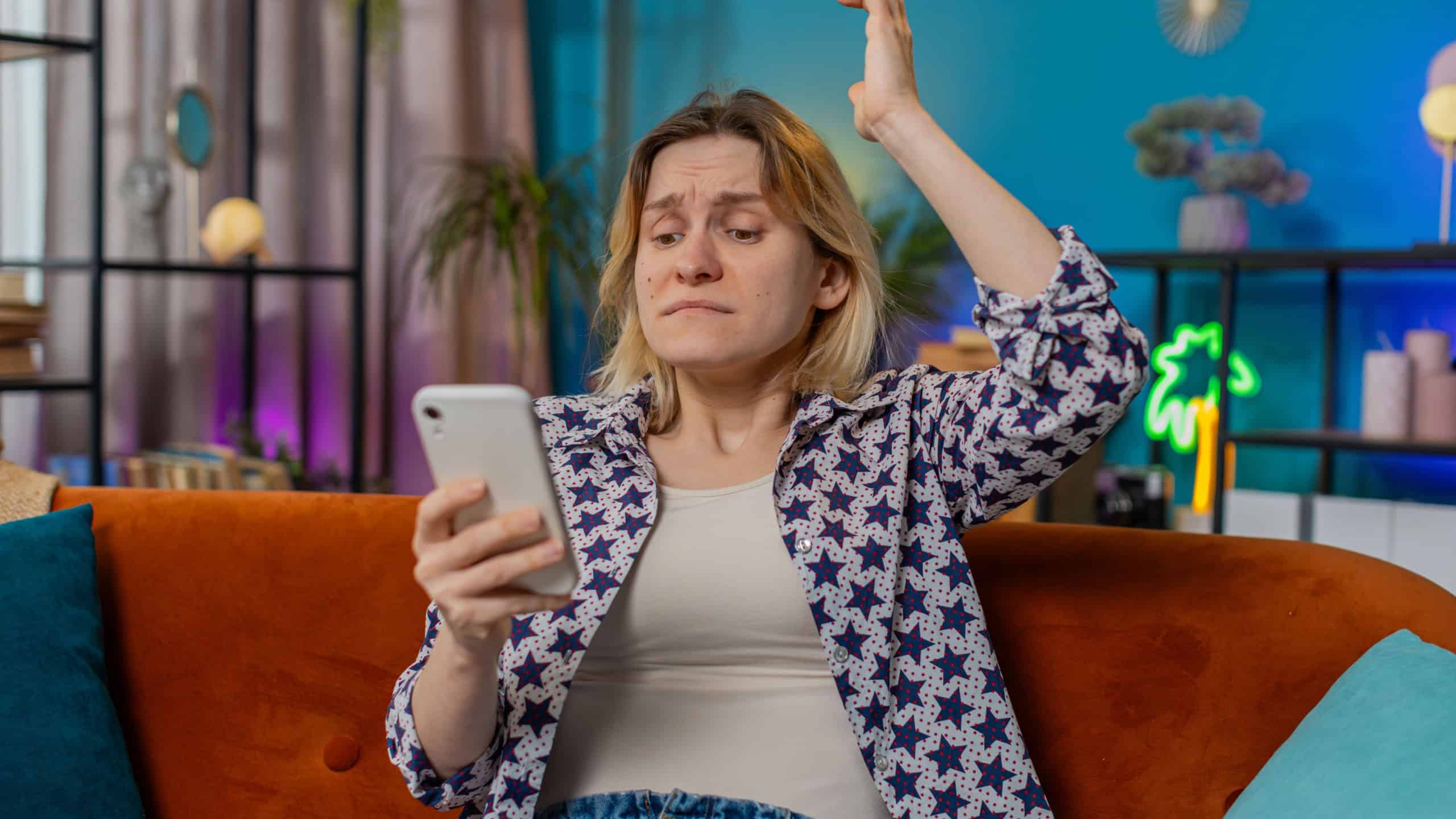 Shocked sad young woman using smartphone reading negative message feels annoyed sitting on sofa couch in living room at home. 