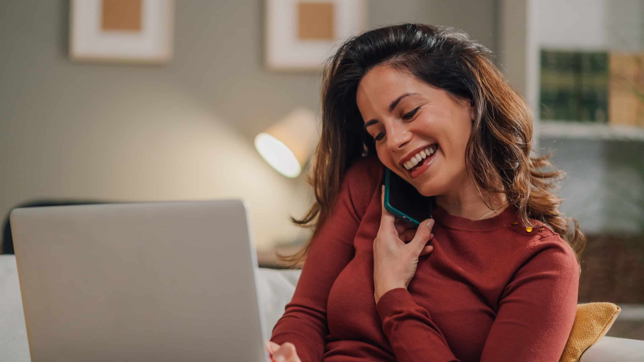 Smiling woman is working from home, comfortably sitting on her sofa while talking on the phone and using her laptop, enjoying a productive and pleasant workday