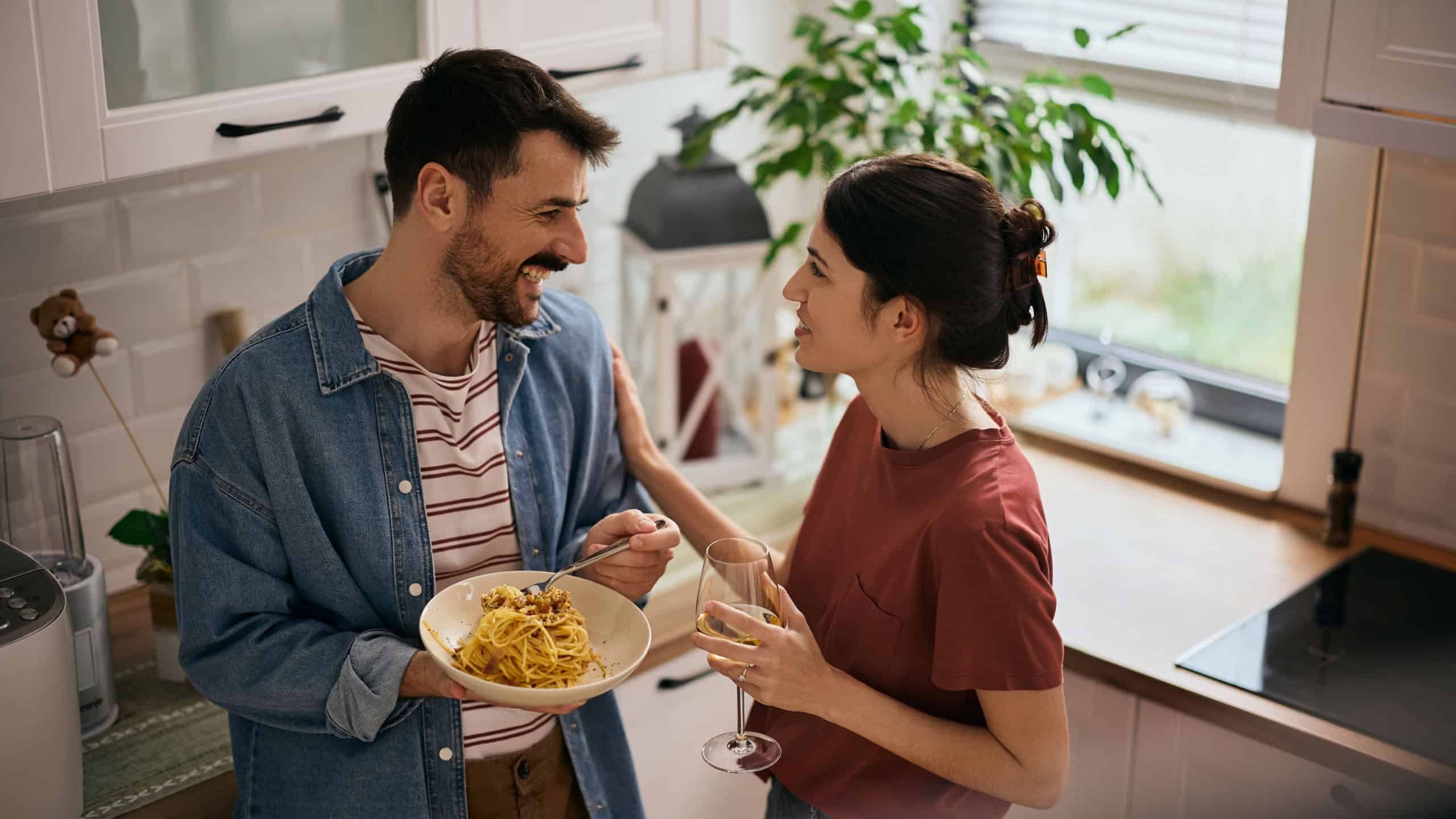 Happy couple talking while drinking wine and eating spaghetti in the kitchen.