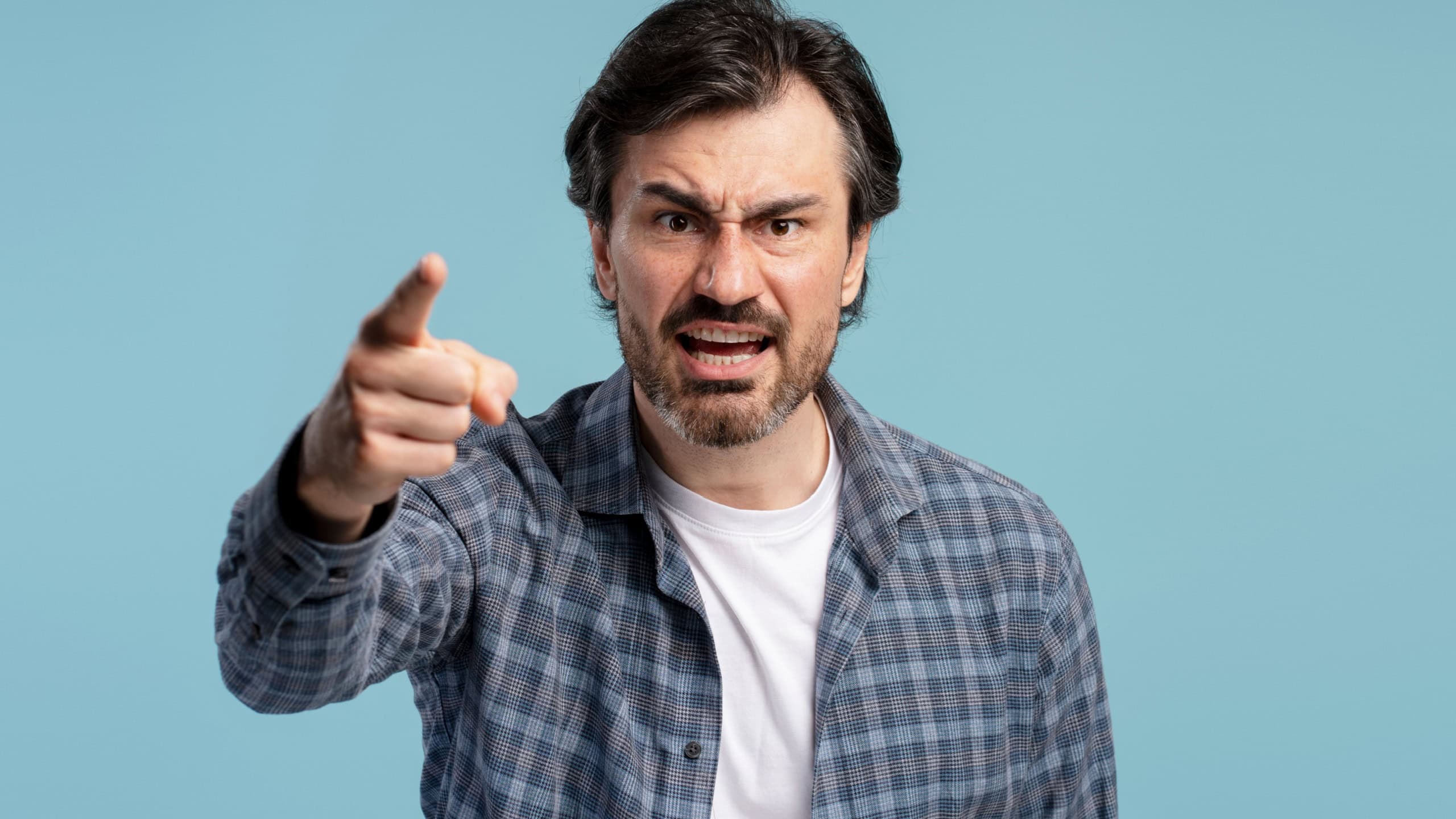 Furious mature man wearing casual clothes pointing finger and shouting on blue studio background, expressing negative emotions like stress, anger, rage and frustration