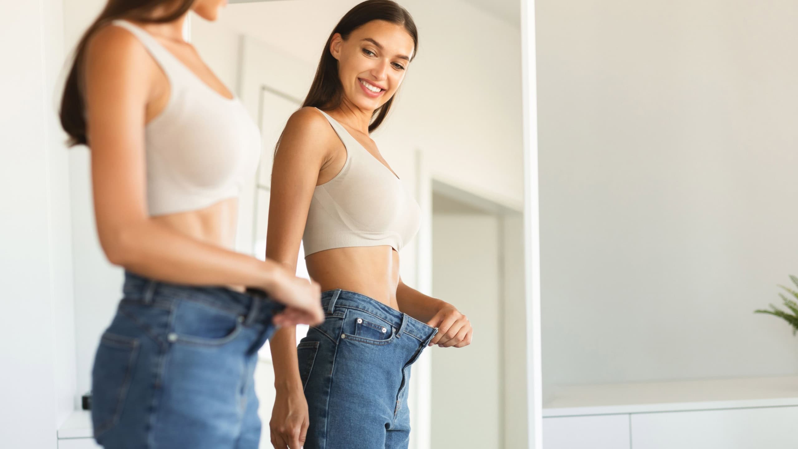 Excited young fit woman losing weight and wearing old too big jeans, woman feeling satisfied with results of her diet and slimming, posing near mirror