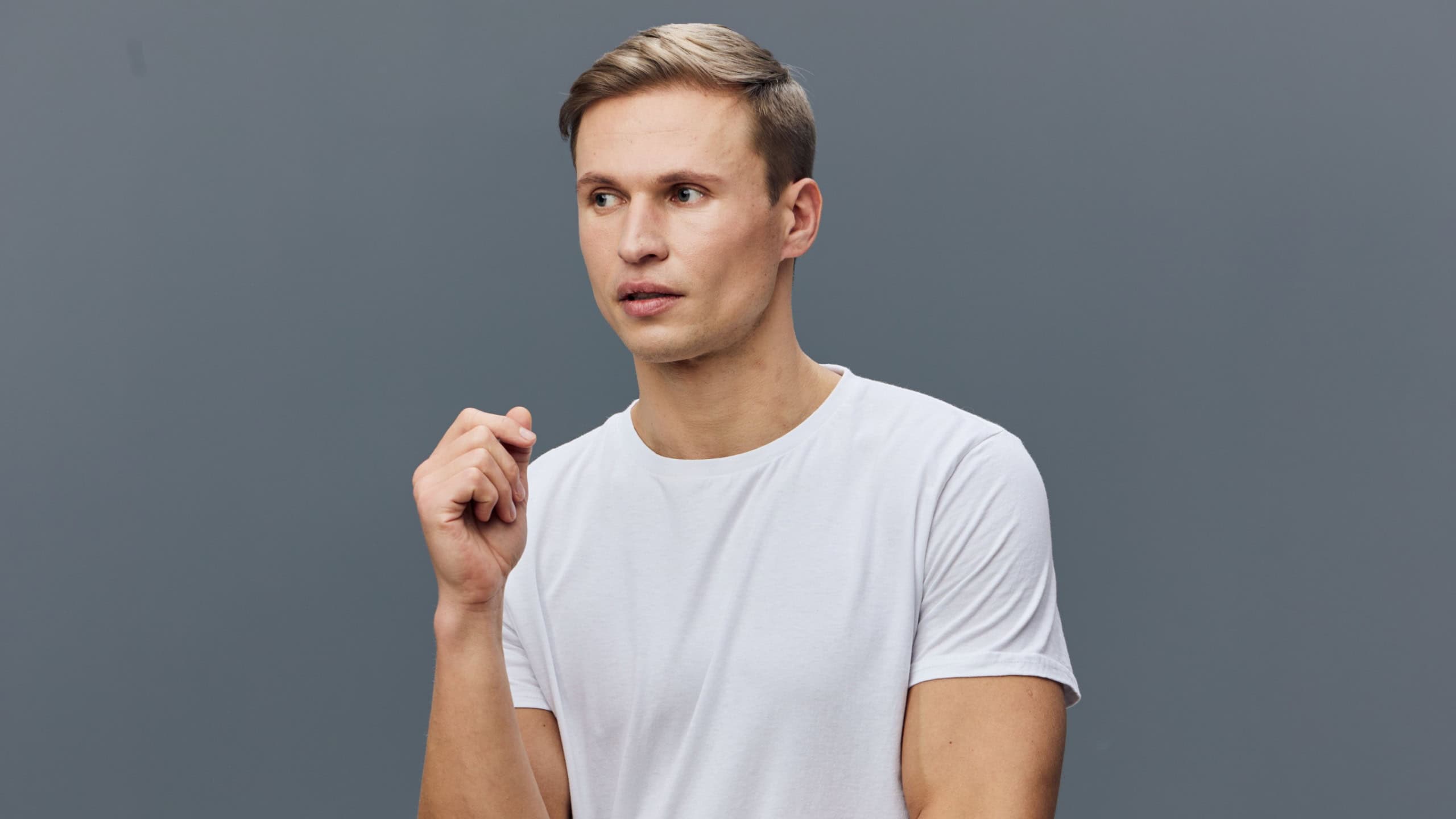 Close-up portrait of a young caucasian man with short hair wearing a plain white t-shirt, looking thoughtful and pensive, isolated on a solid gray background, studio shot, lifestyle concept