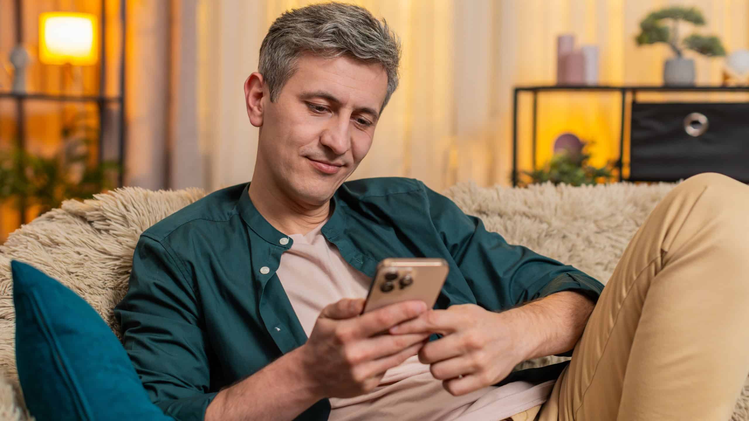 Young man at home sits on sofa smiling warmly while looking at old family photos on smartphone. Caucasian guy enjoys nostalgic feelings and soft memories, thinking about relatives and childhood days