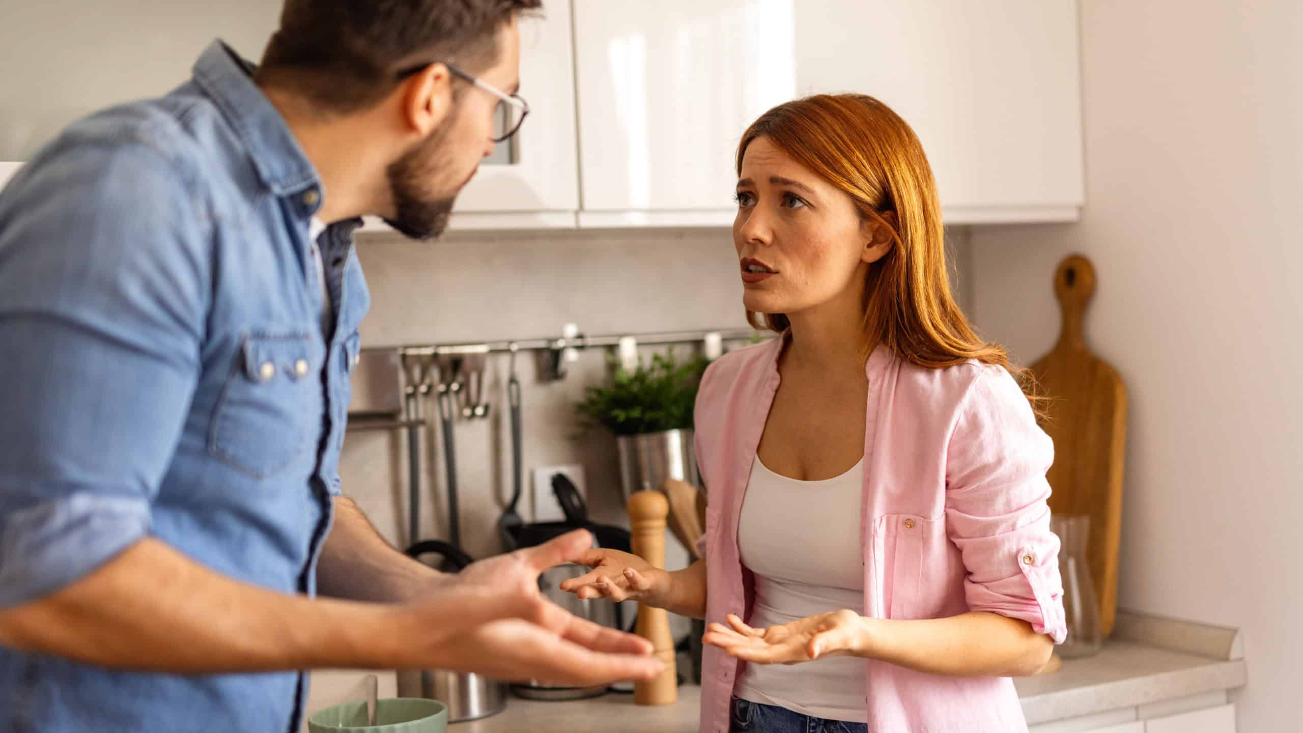 Intense disagreement between a couple in their kitchen. The man raises his hand in a defensive manner. Both display strong emotions, reflecting a challenging moment in their relationship
