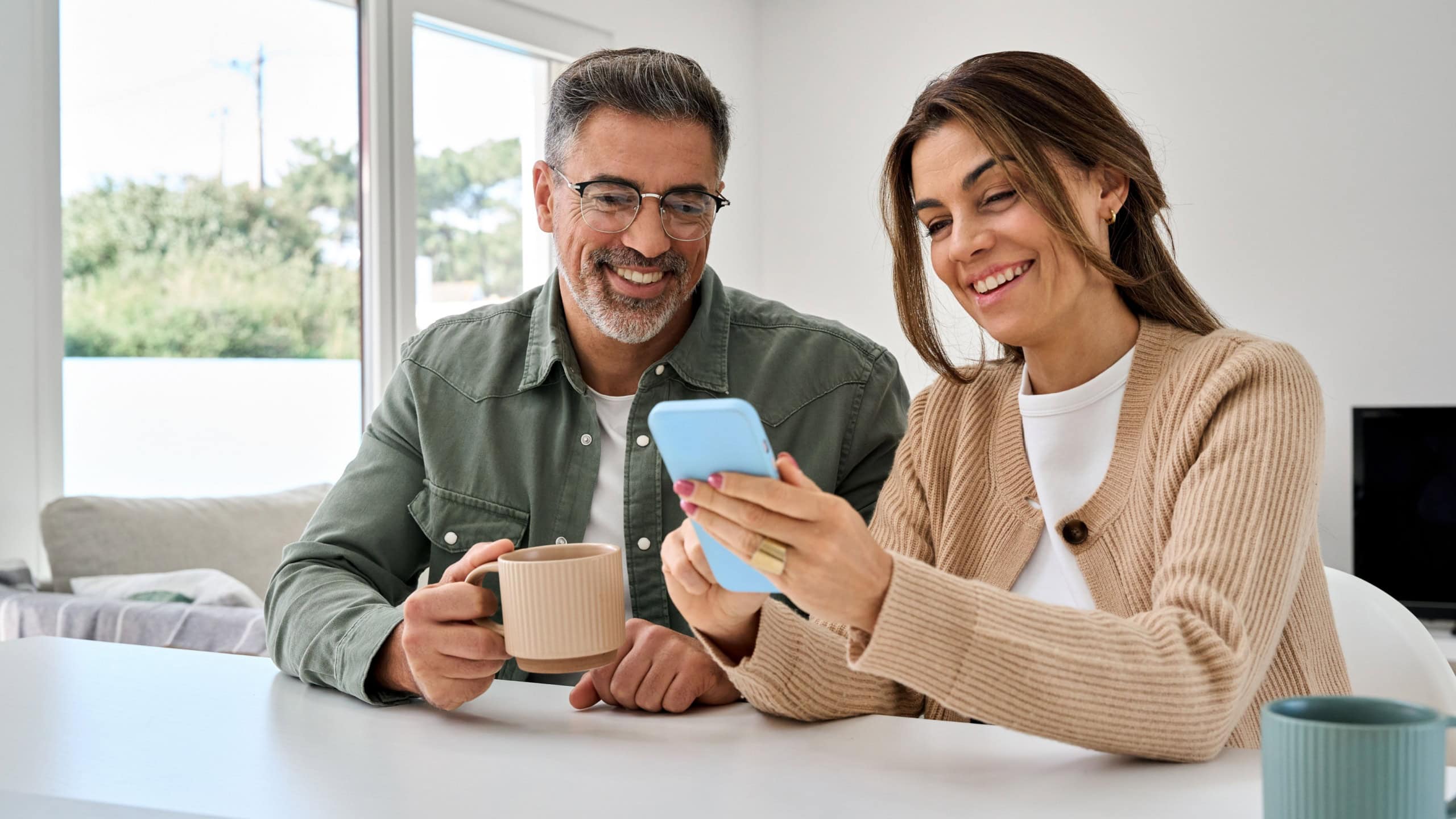 Smiling mature older man and woman holding cellphone browsing internet, texting message on mobile cell phone technology. Middle aged happy couple using smartphone relaxing at living room table at home