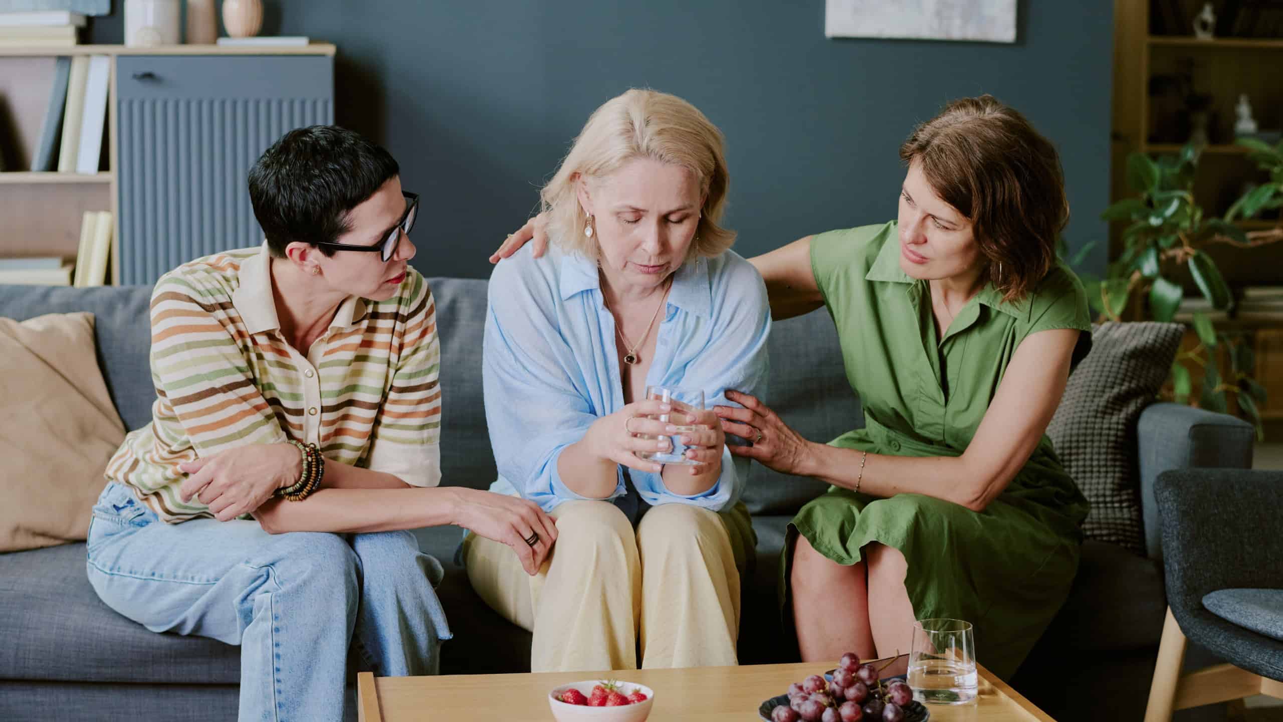 Middle aged Caucasian woman sitting on sofa holding glass of water looking down, supported by two caring girlfriends comforting her with gentle touch, bowl of fruit on table
