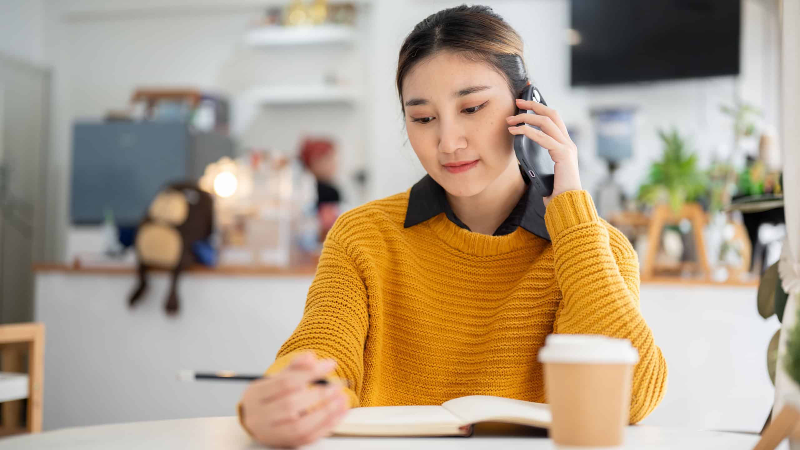Young asian woman holding pencil over notebook and making a phone call while sitting at cafe's table. Working or Study, Chilling Outside, Urban life.
