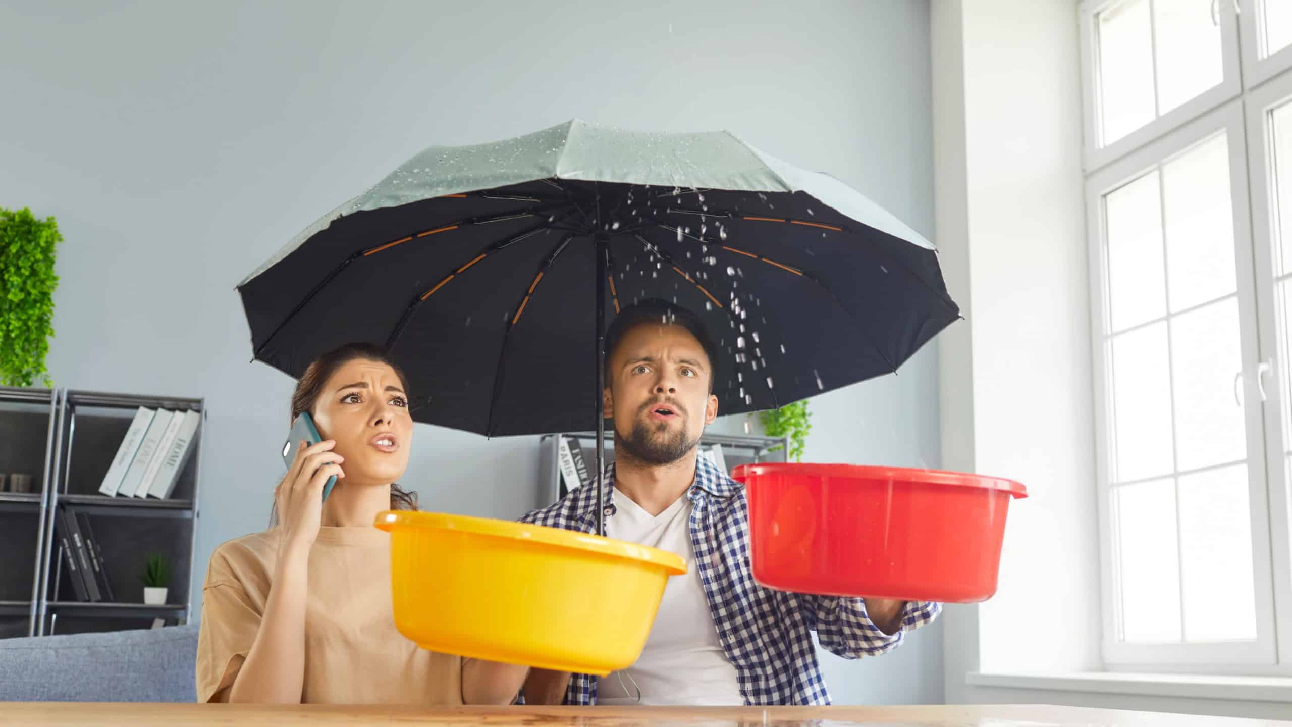 Young stressed couple sits at table under open umbrella, collecting water drips in buckets, experiencing household issue, roof leaks or plumbing problems, calling to home repair services