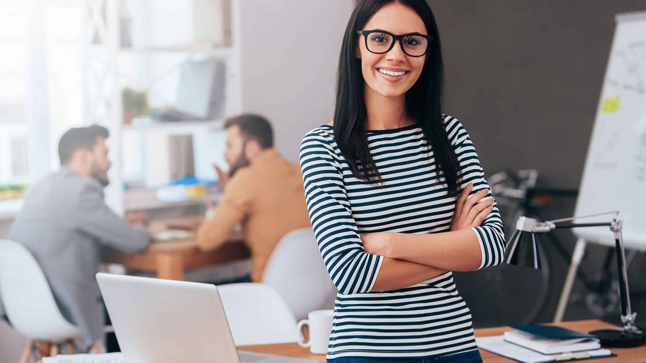Confident and successful leader. Confident young woman keeping arms crossed and looking at camera with smile while her colleagues working in the background