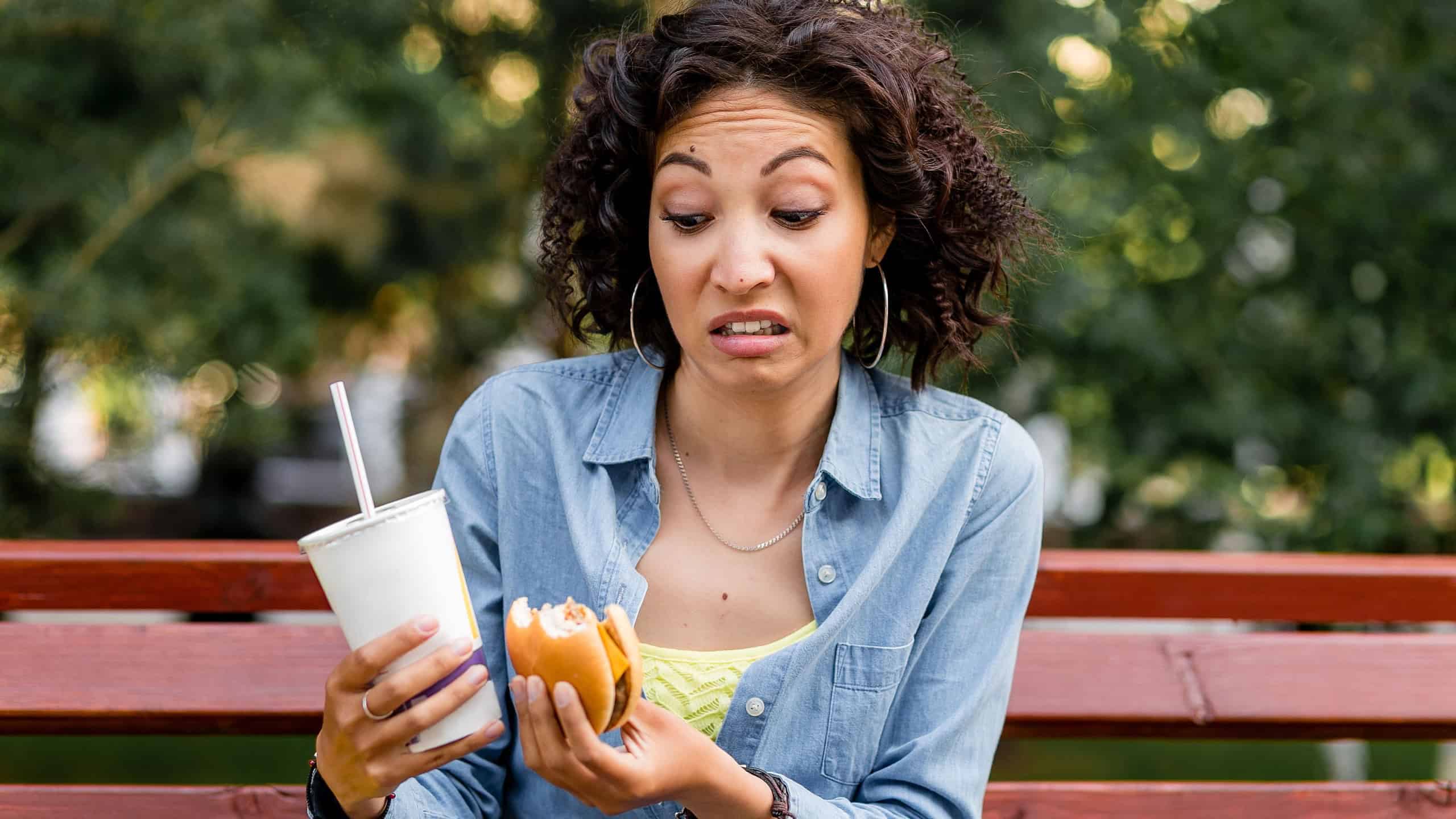 Outdoor portrait of woman looking at fast food hamburger and soda with disgusting emotion