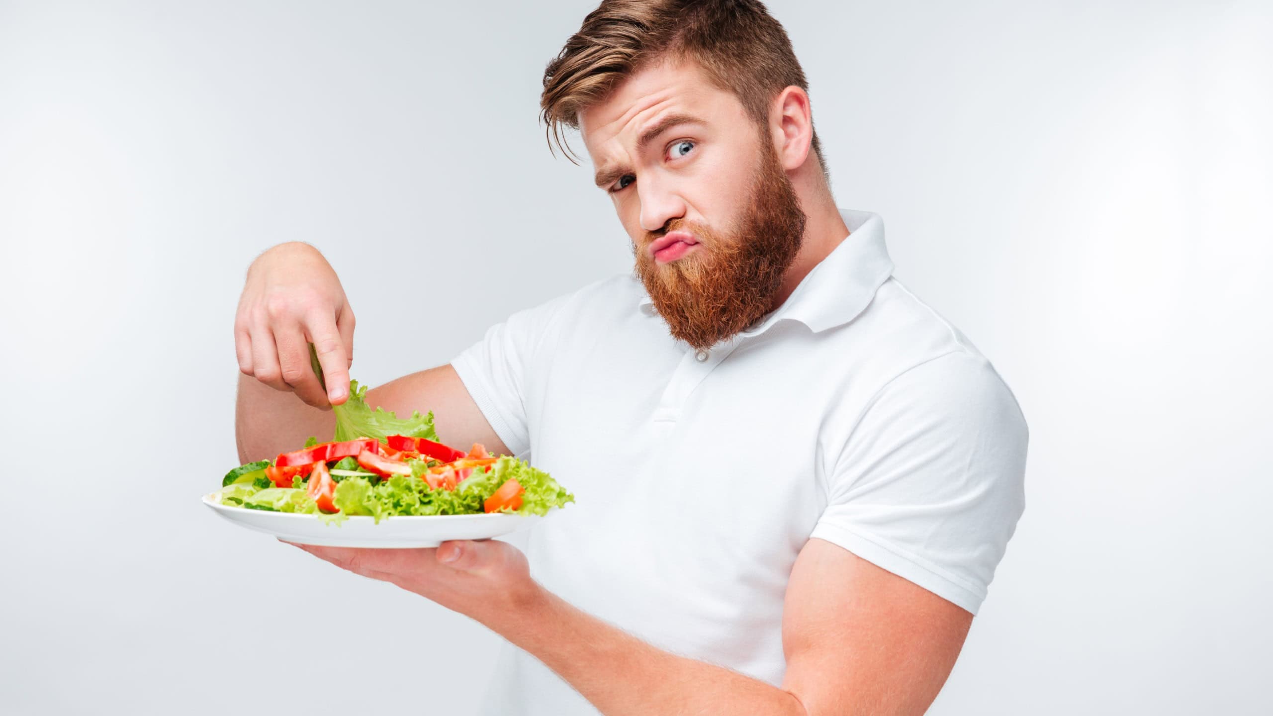 Confused bearded man holding lettuce and holding plate with salad isolated on white background