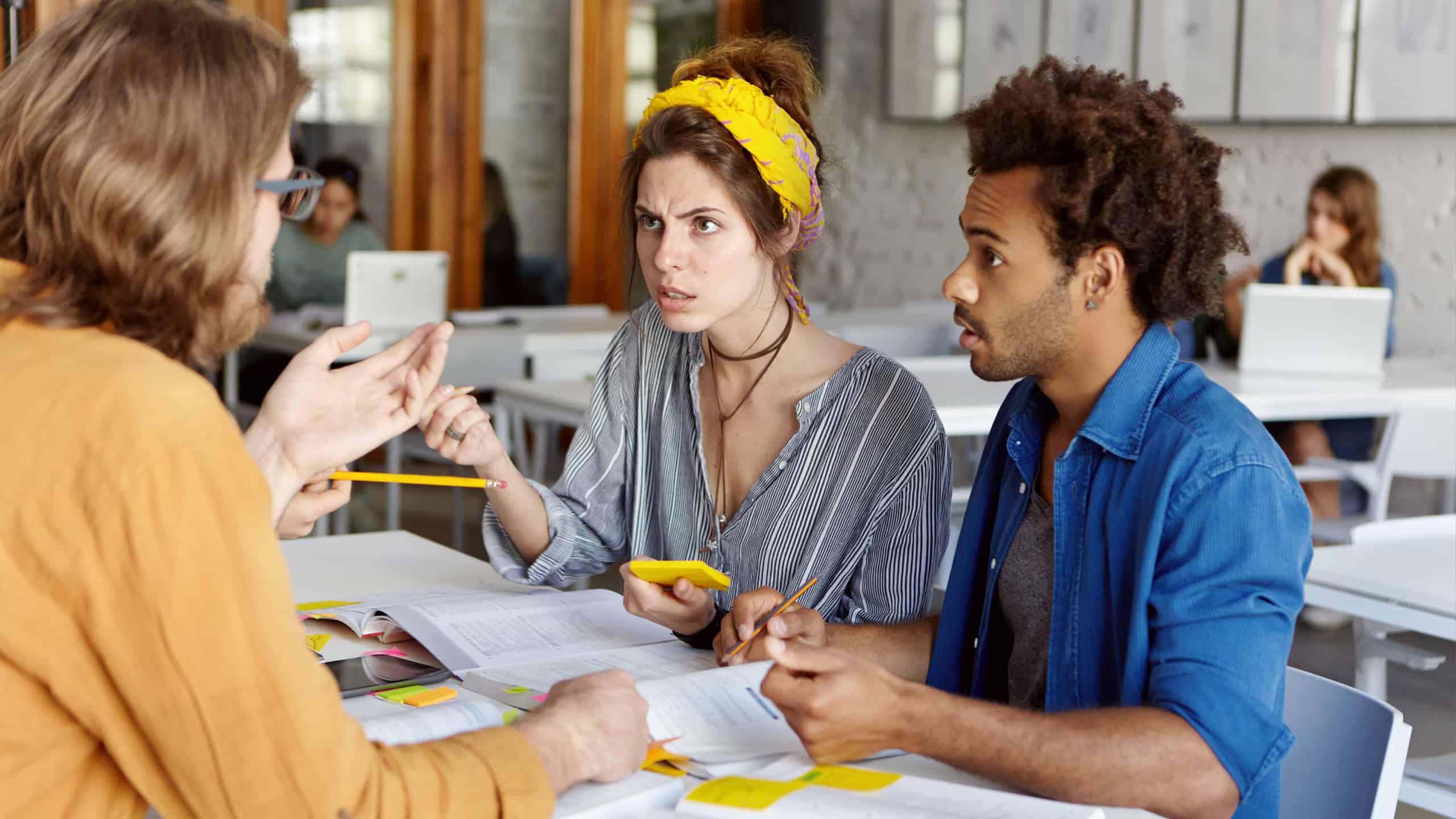 Clueless Caucasian young woman and Afro-American man students having confused expressions, trying to understand what their teacher in glasses is talking about during Spanish class at coffee shop