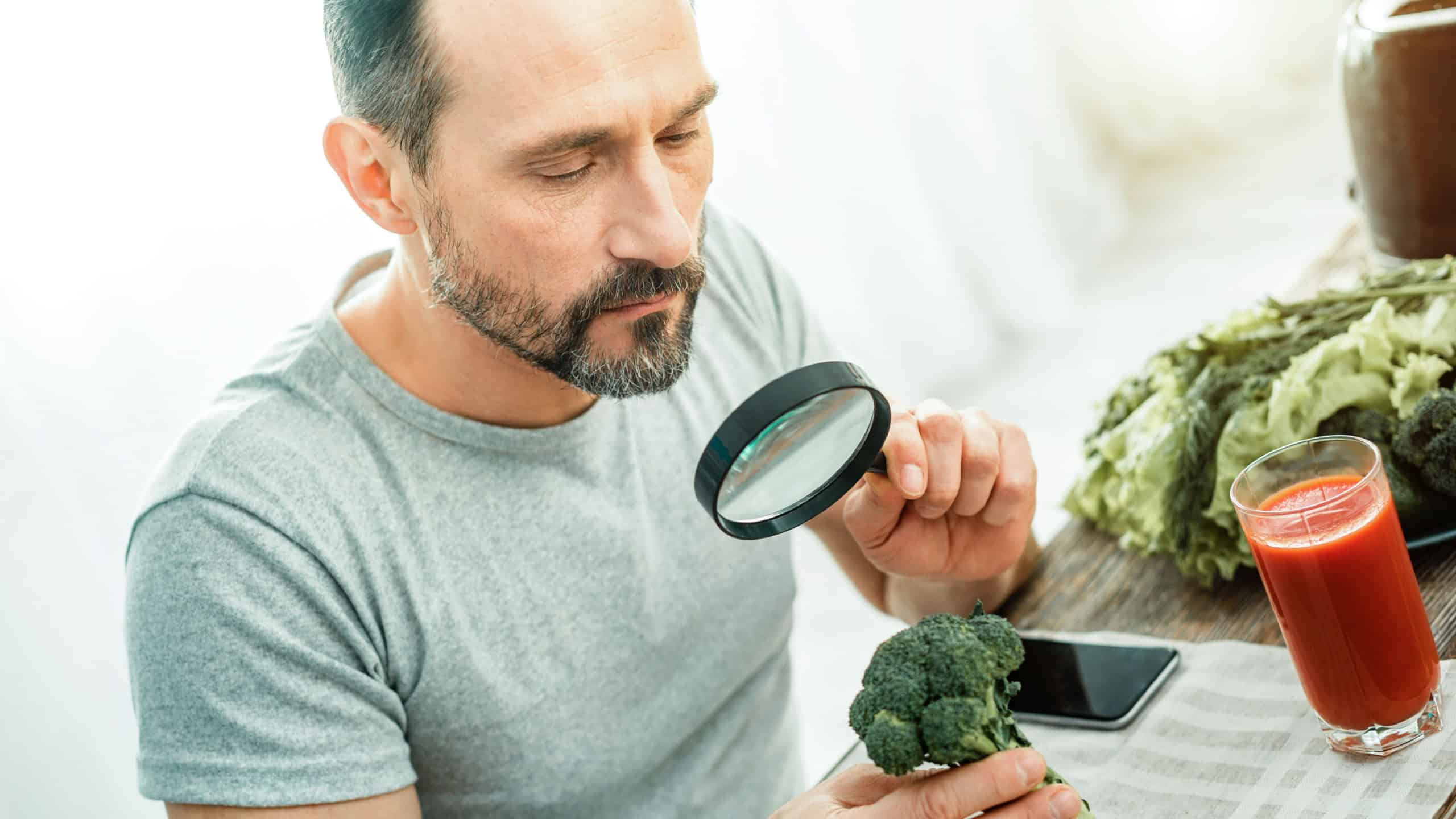 Strange plant. Concentrated occupied calm man sitting by the table holding the and magnifying glass examining a broccoli.