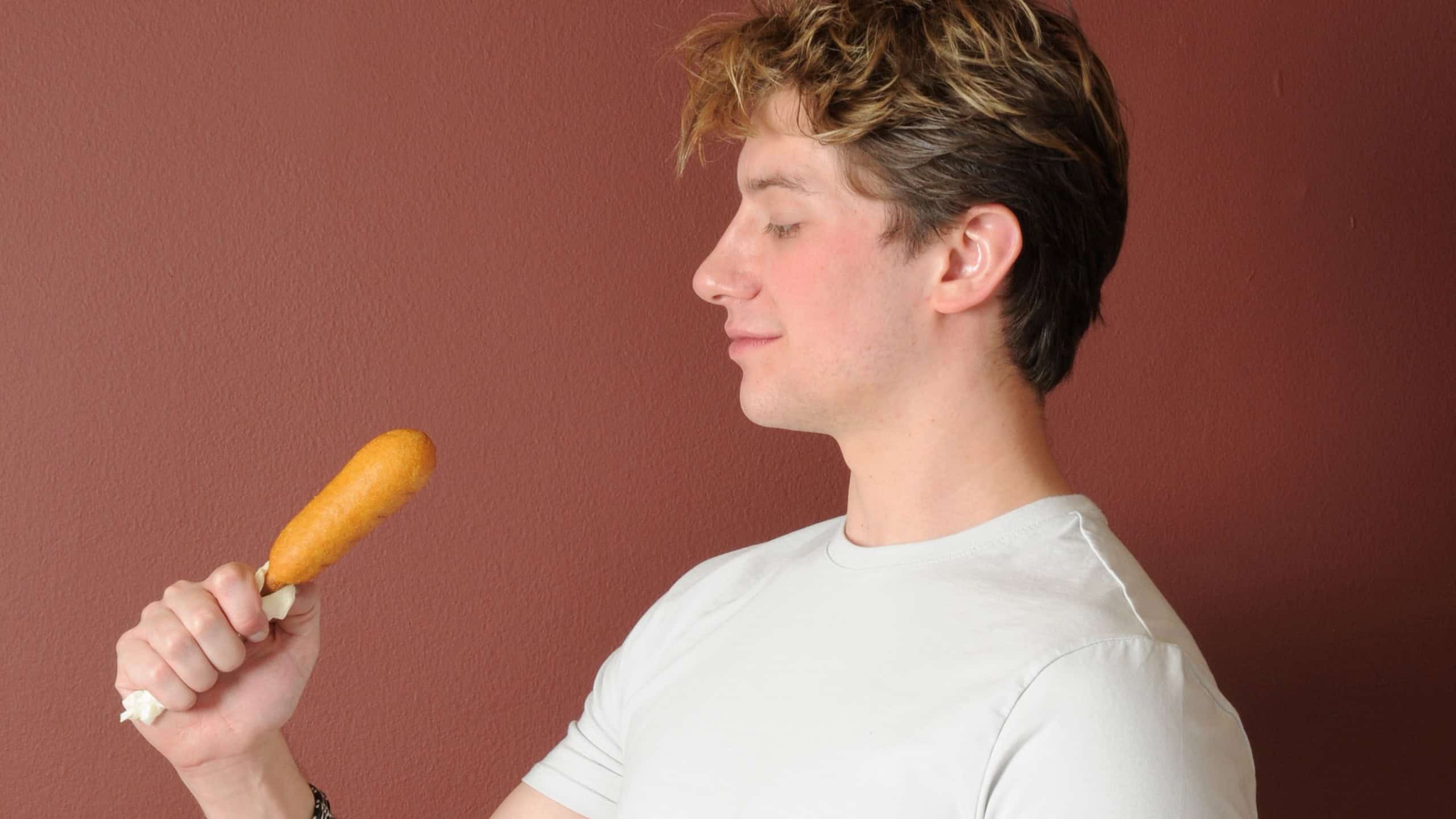 A handsome young blond man about to bite a corn dog