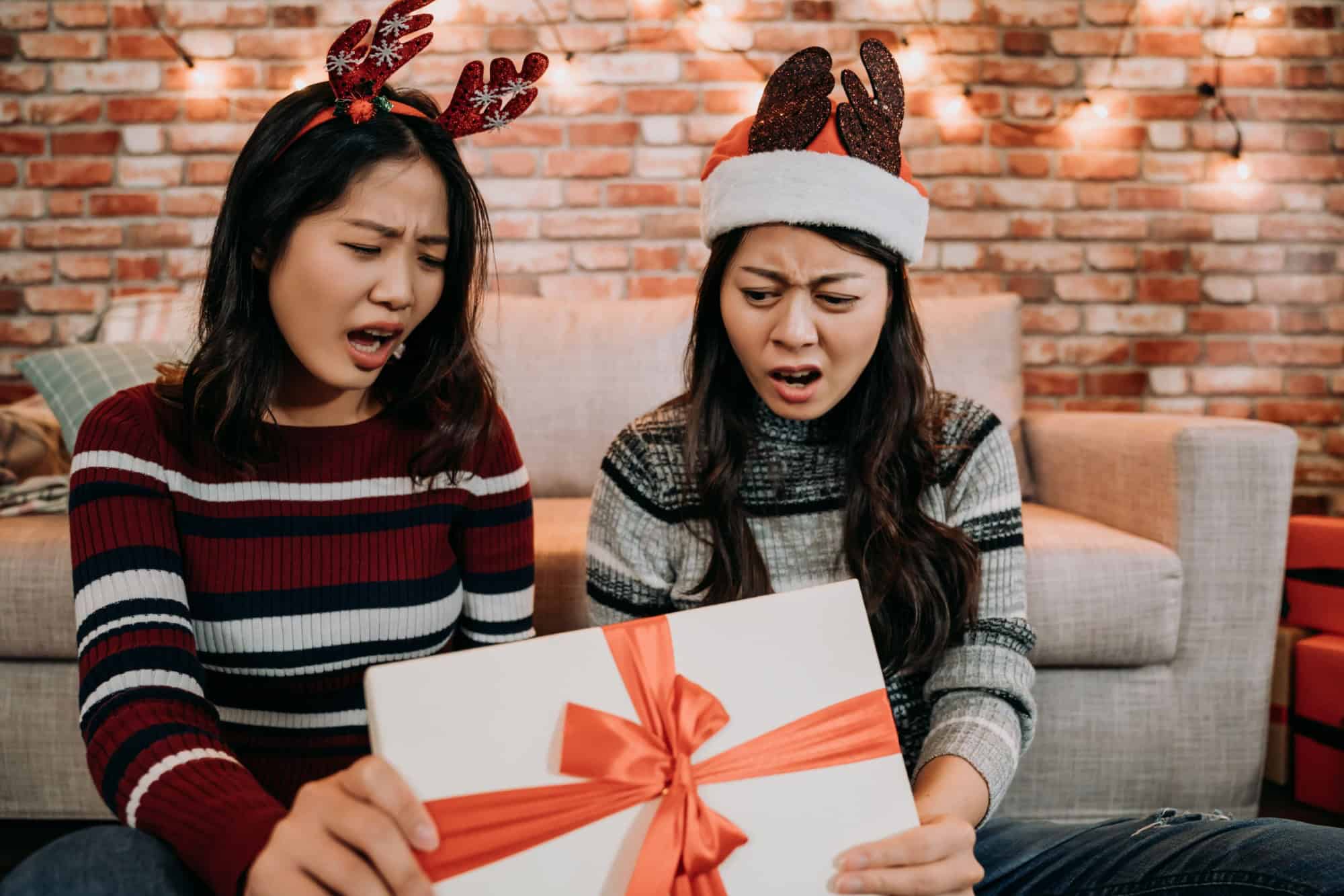 sisters received a terrible present from online shopping. young girls open gift box day at home feeling displeased. grumpy girls in santa hats buying clothes from christmas sale.