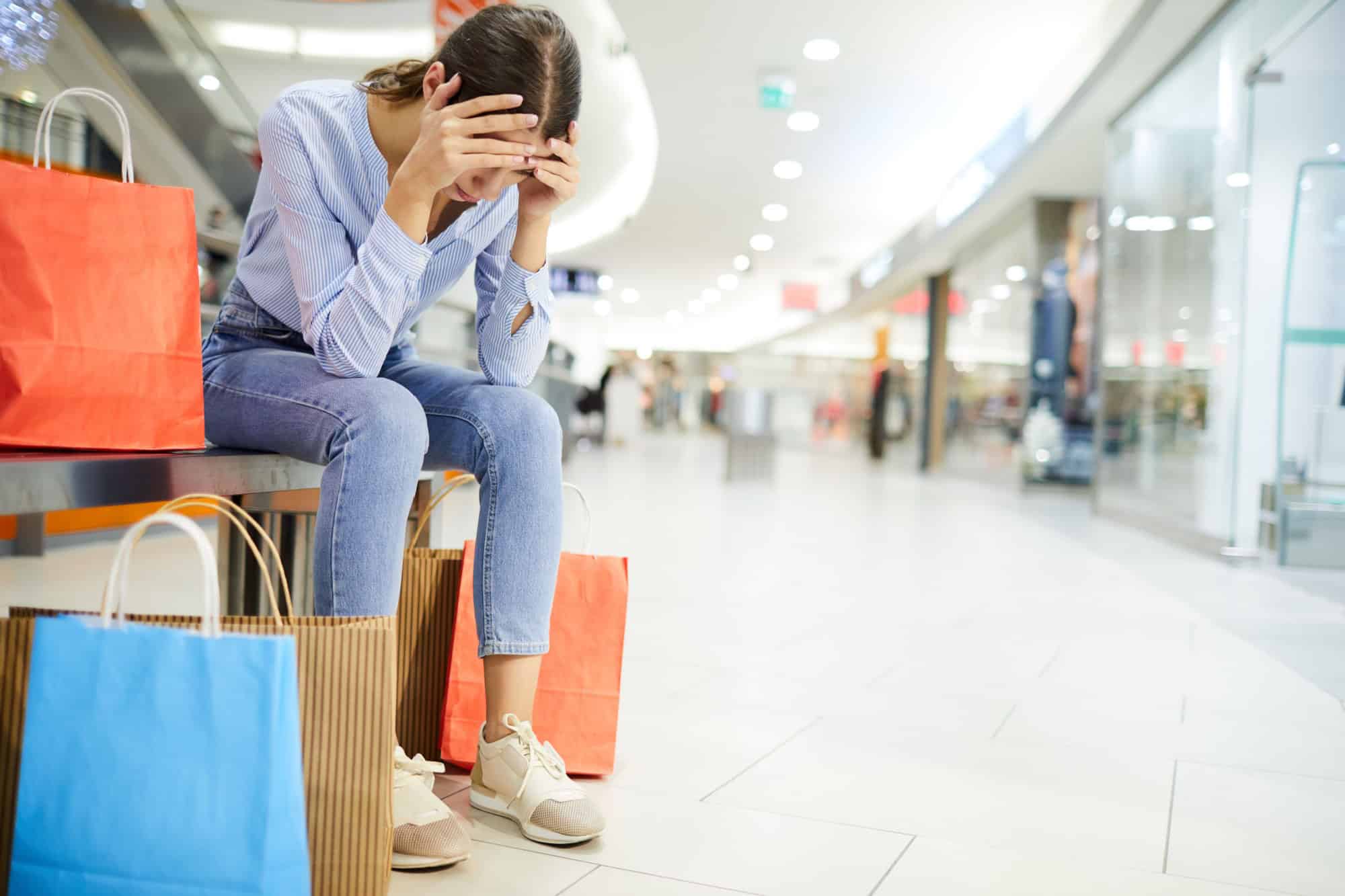 Tired shopper holding by head while sitting on bench among paperbags with purchases in the mall