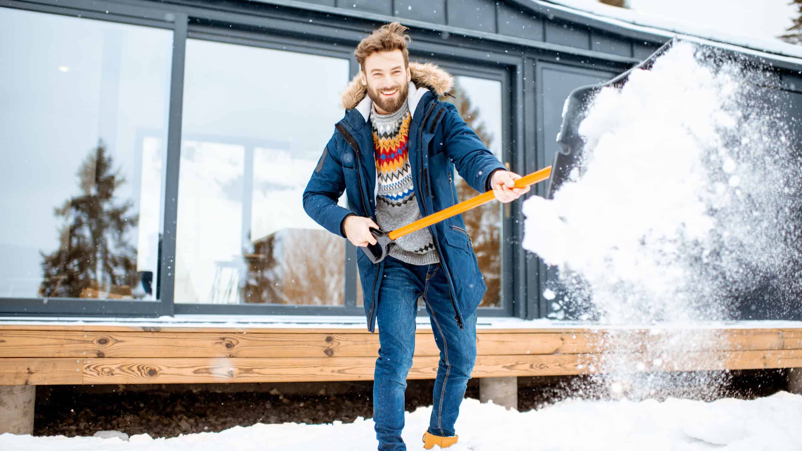 Handsome man in winter clothes cleaning snow with a shovel near the modern house in the mountains