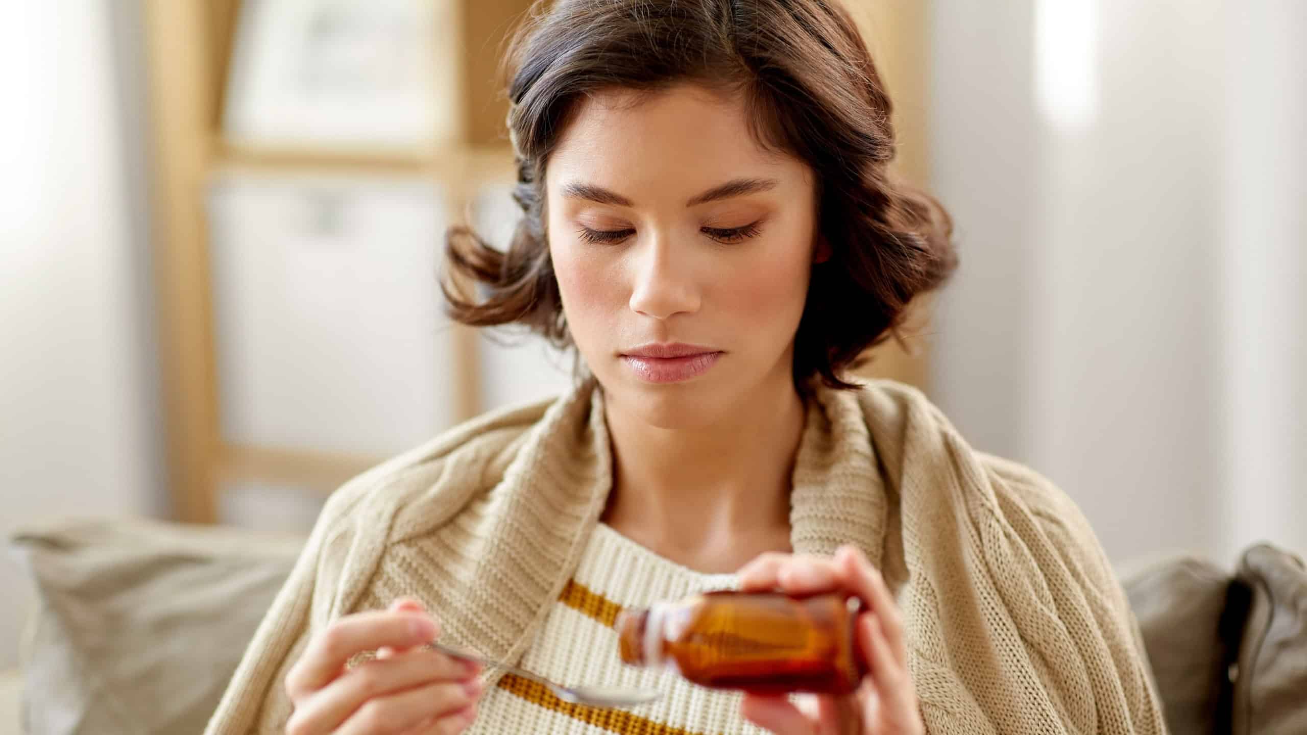 People, health and medicine concept - sad sick young woman in blanket pouring antipyretic or cough syrup from bottle to spoon at home