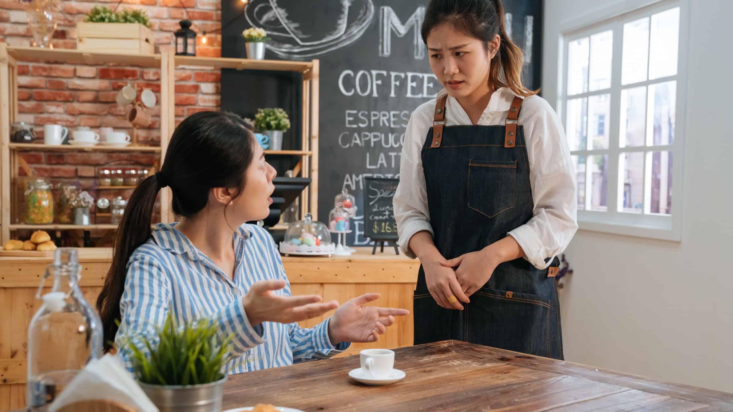 Upset young businesswoman complain about cup of hot coffee. woman customer hold it in hands and point to order while look at waitress. lady client complaining meal to waitress in cafe store.